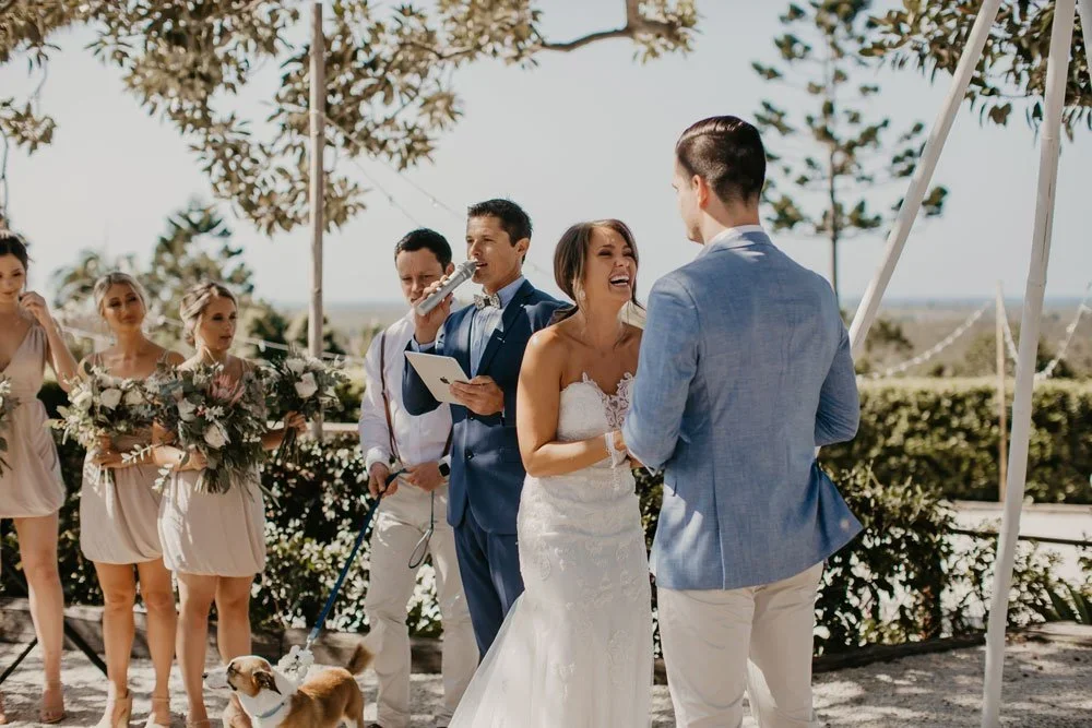 A wedding ceremony outdoors with a bride and groom exchanging vows, surrounded by bridesmaids and groomsmen, with a officiant holding a microphone, a dog, and trees in the background.