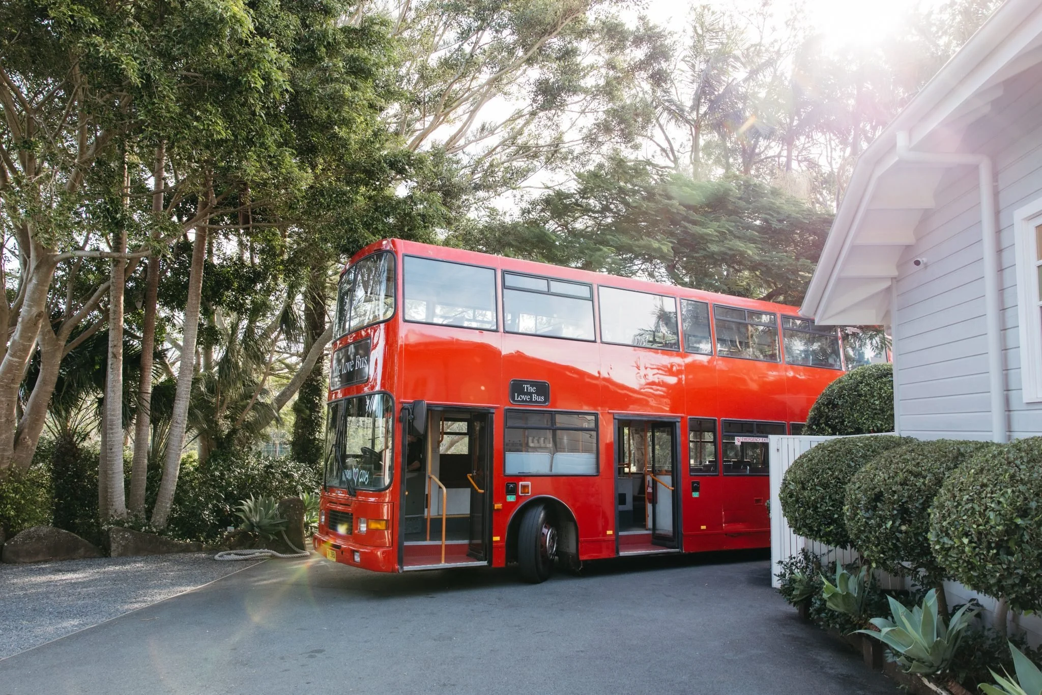 A red double-decker bus parked outdoors near a white house with white siding, surrounded by trees and bushes, with sunlight filtering through the trees.