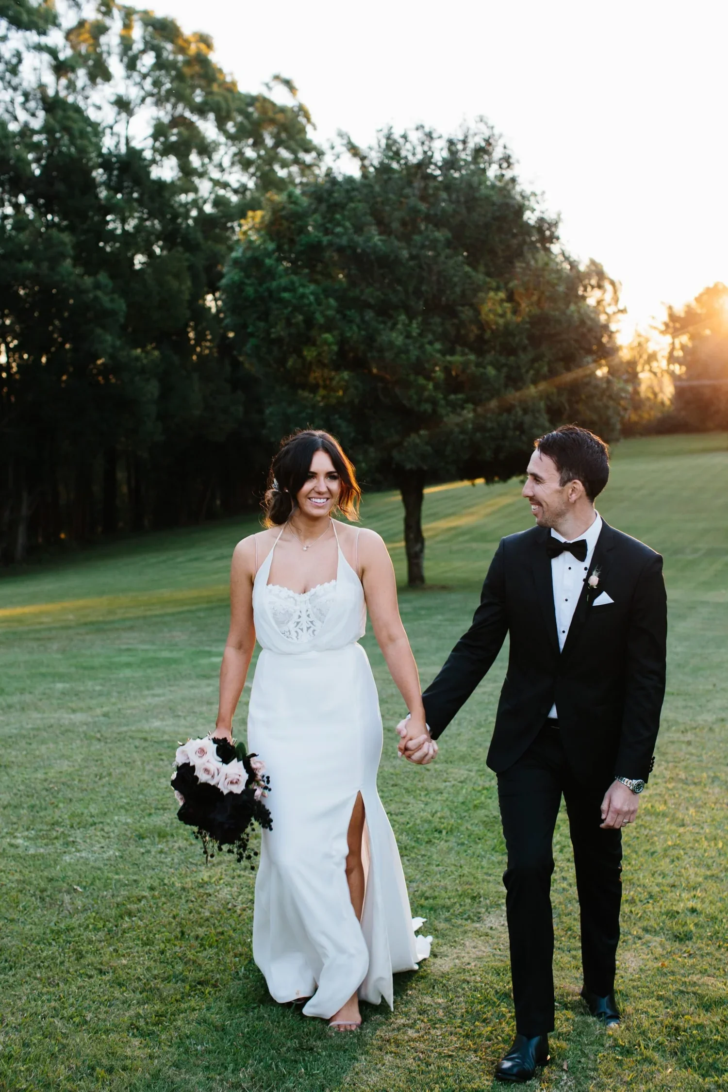 A bride and groom walking hand in hand on a grassy field at sunset, smiling at each other. The bride wears a white wedding dress and holds a bouquet of flowers, while the groom wears a black tuxedo with a bow tie.