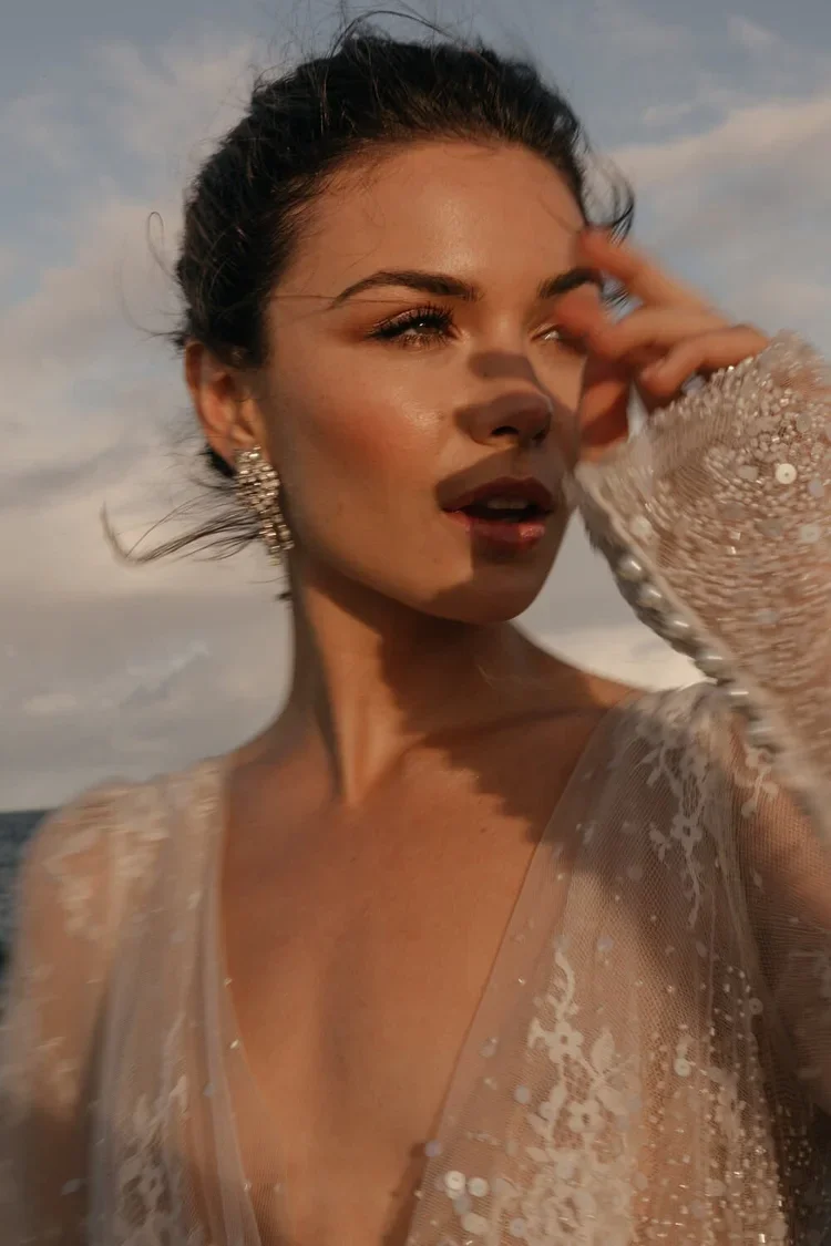 A woman with dark hair tied back, wearing large earrings and a sheer, embellished dress, is touching her face with her hand while looking into the distance outdoors during sunset.