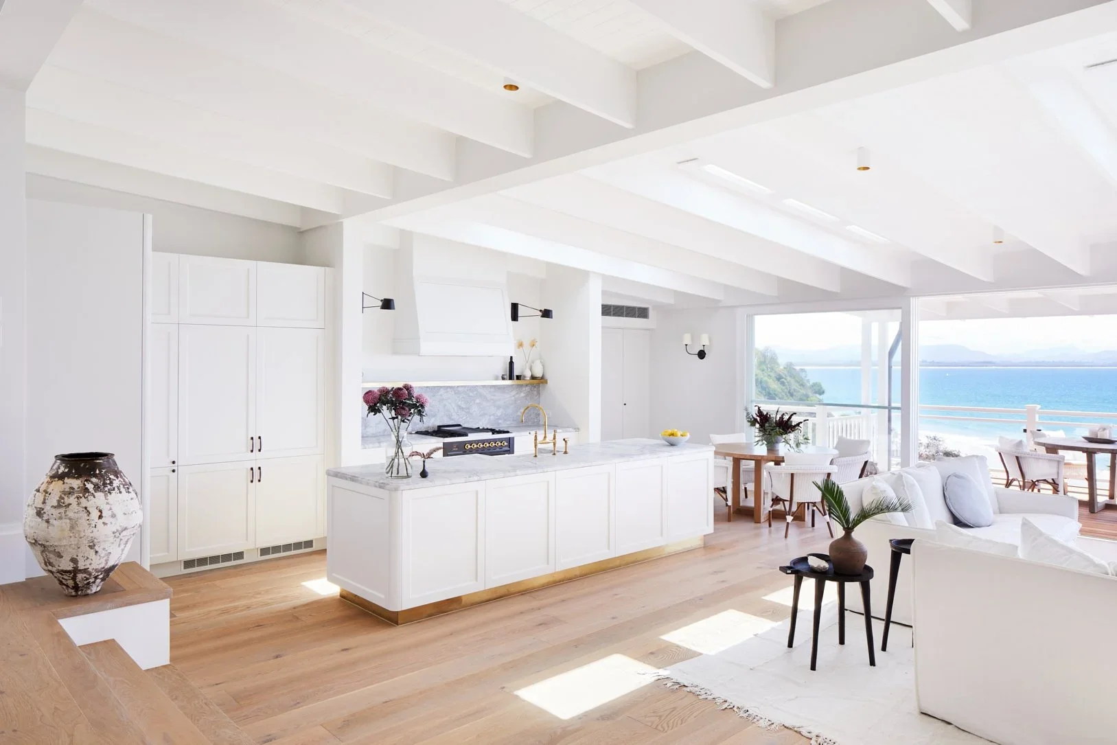 A bright, modern open-plan living space with a white kitchen island, light wood flooring, large windows overlooking a body of water, and white furniture, including a sofa and dining table.