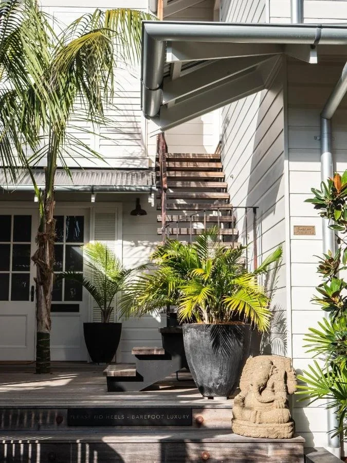 Exterior view of a house with stairs leading up to an upper floor, potted plants on a porch, a stone sculpture, and tropical trees, in bright daylight.