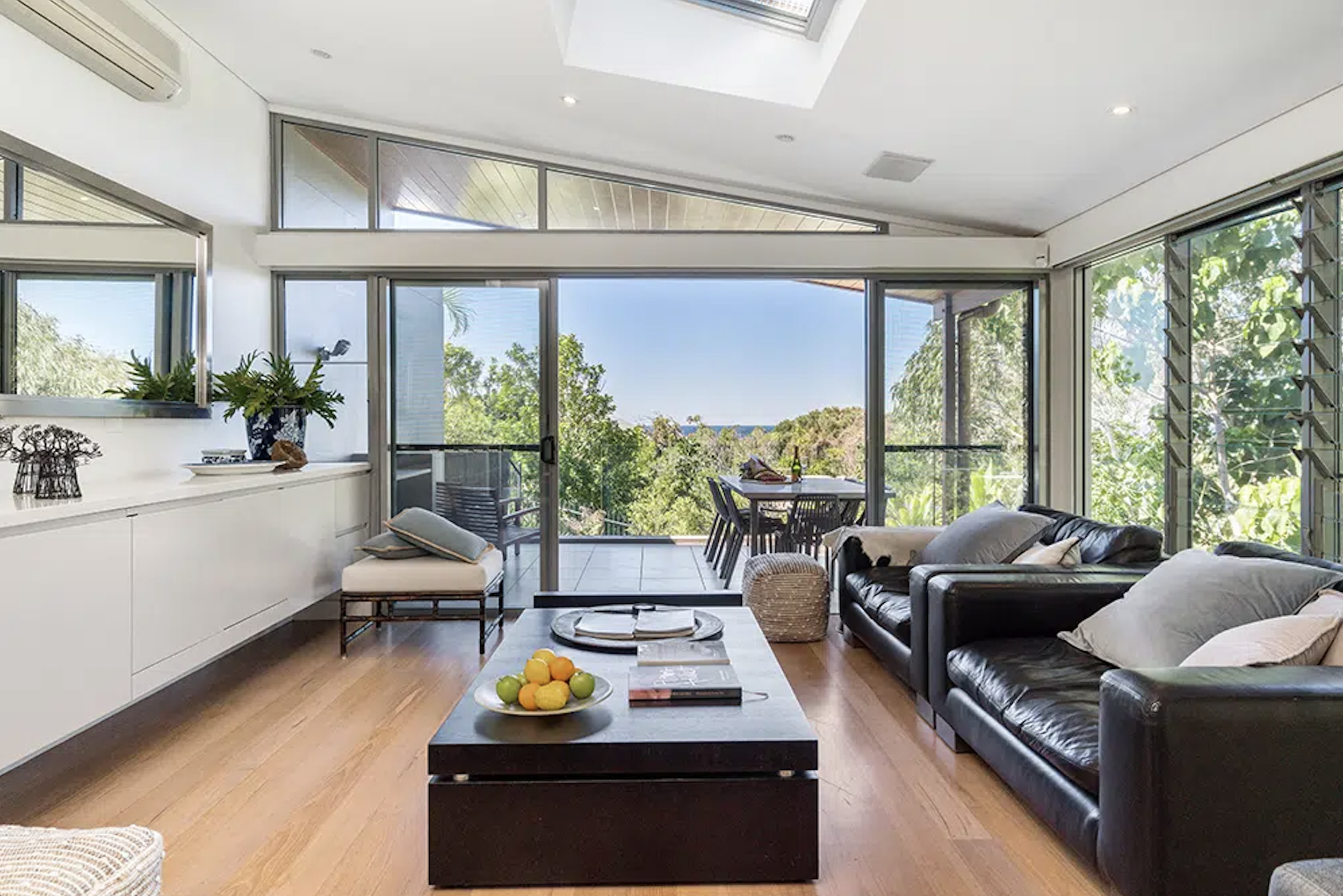Living room with large glass doors leading to a balcony, overlooking trees and the ocean. Furniture includes a black leather sofa with pillows, a wooden coffee table with fruit and books, and a white sideboard decorated with plants and sculptures. The ceiling has a skylight, and the floor is wooden.