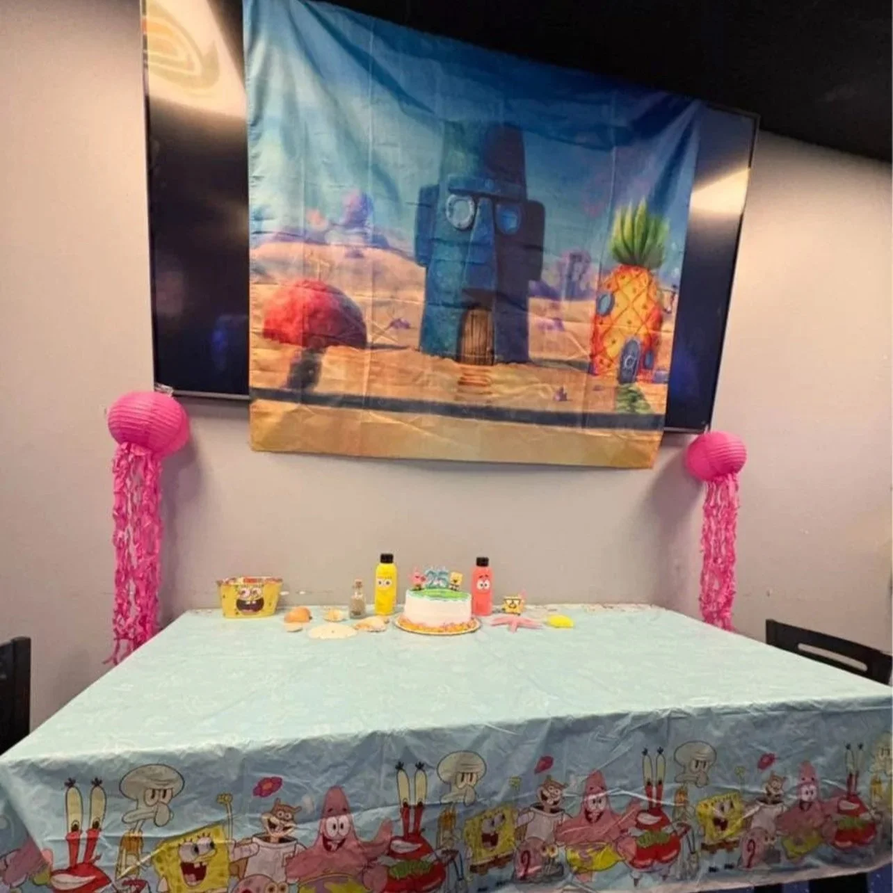 Decorated table for a children's birthday party with SpongeBob SquarePants theme. There are pink paper lanterns with streamers hanging from them on each side. The table has a SpongeBob-themed tablecloth, and various party decorations including a small cake, bottles, and seashell souvenirs. A colorful image of SpongeBob's pineapple house and underwater scene is displayed on a large poster hanging behind the table.