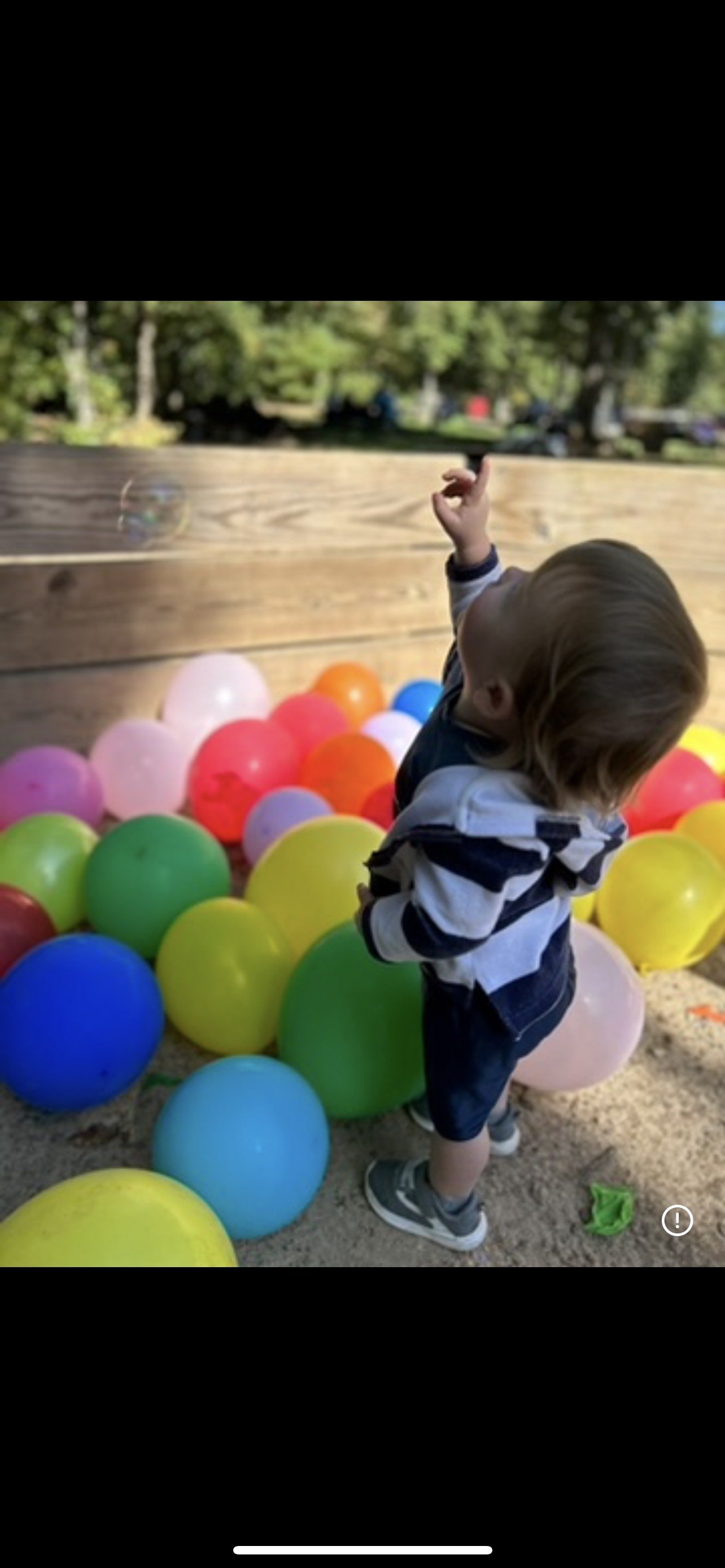 A young child with blonde hair wearing a black and white striped hoodie, shorts, and sneakers, standing among colorful balloons outdoors on a sandy surface with trees in the background, reaching towards a floating soap bubble.