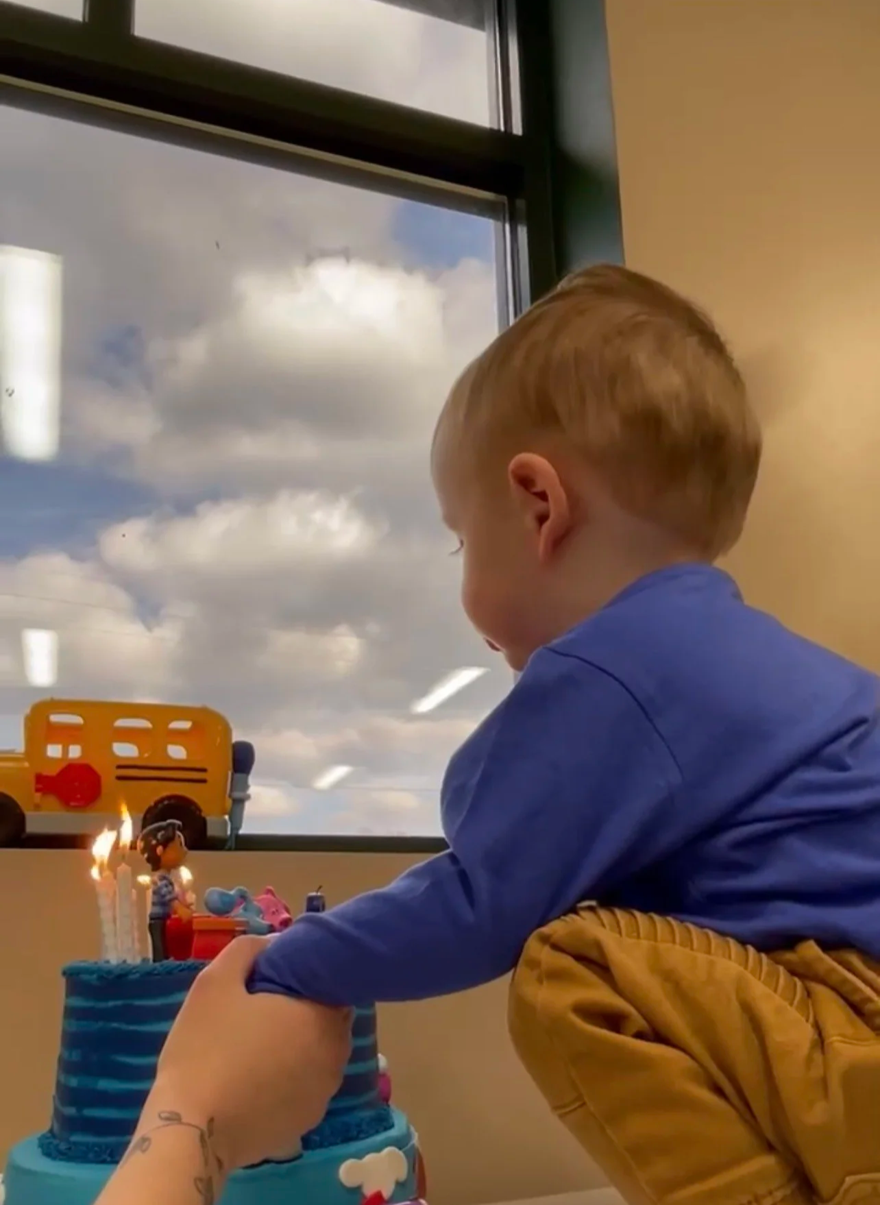 A young boy celebrates his birthday with a blue cake that has candles and characters on top, while an adult helps him light the candles. They are indoors with large windows showing a cloudy sky outside.