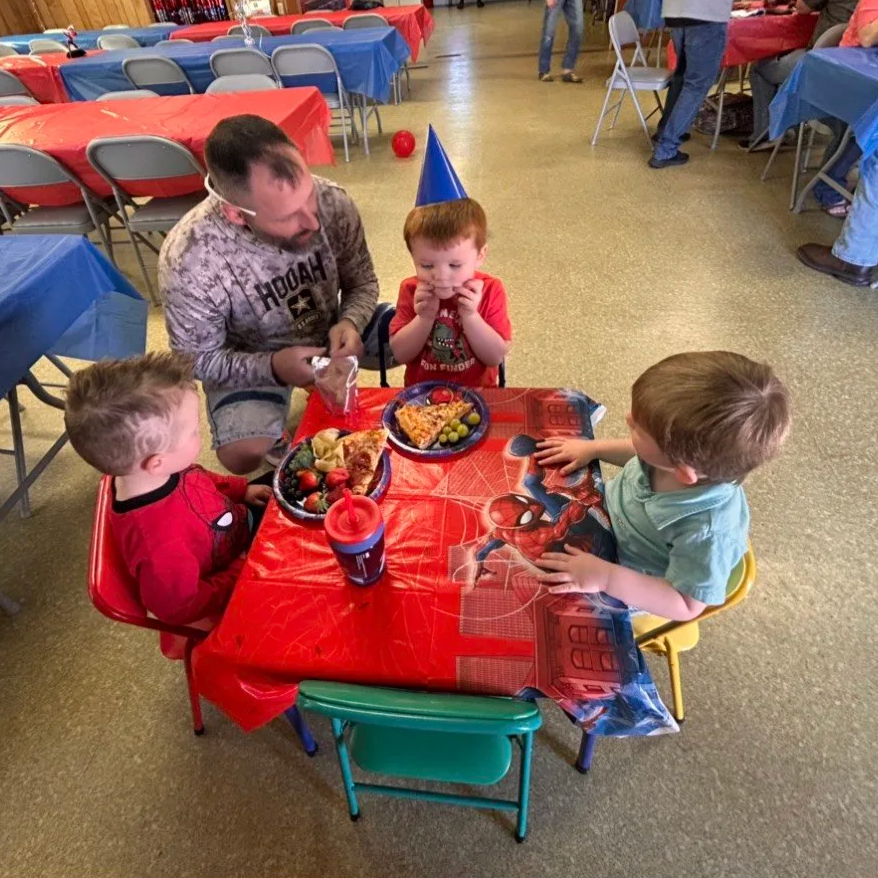 Children celebrating birthday at a party with a Spiderman-themed tablecloth, wearing party hats, with plates of pizza, grapes, and strawberries, and a red ball on the floor in the background.