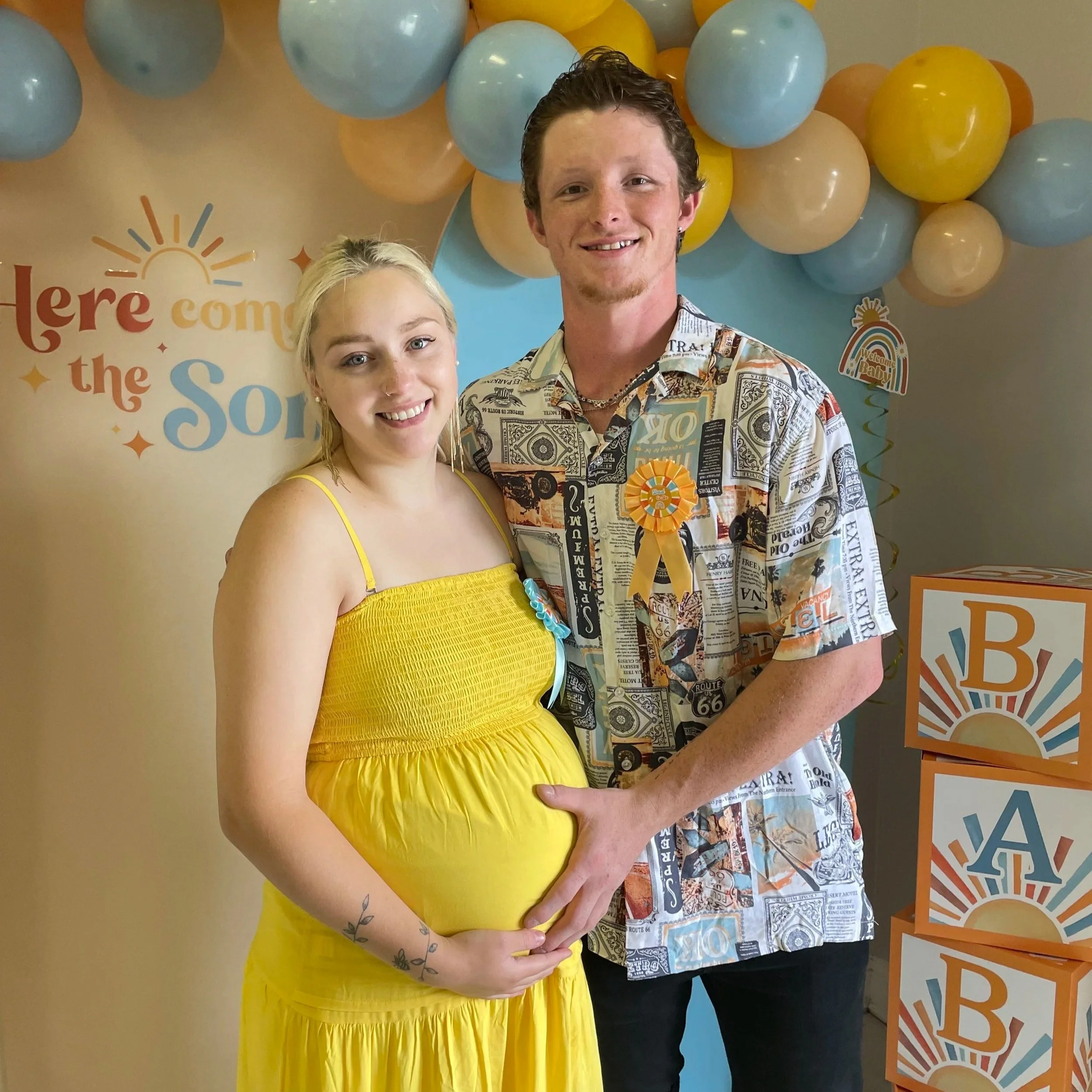 A pregnant woman in a yellow dress and a man in a patterned Hawaiian shirt stand together smiling, with decorations including balloons, blocks spelling 'BABY', and a sign that reads 'Here comes the Sun' in the background.