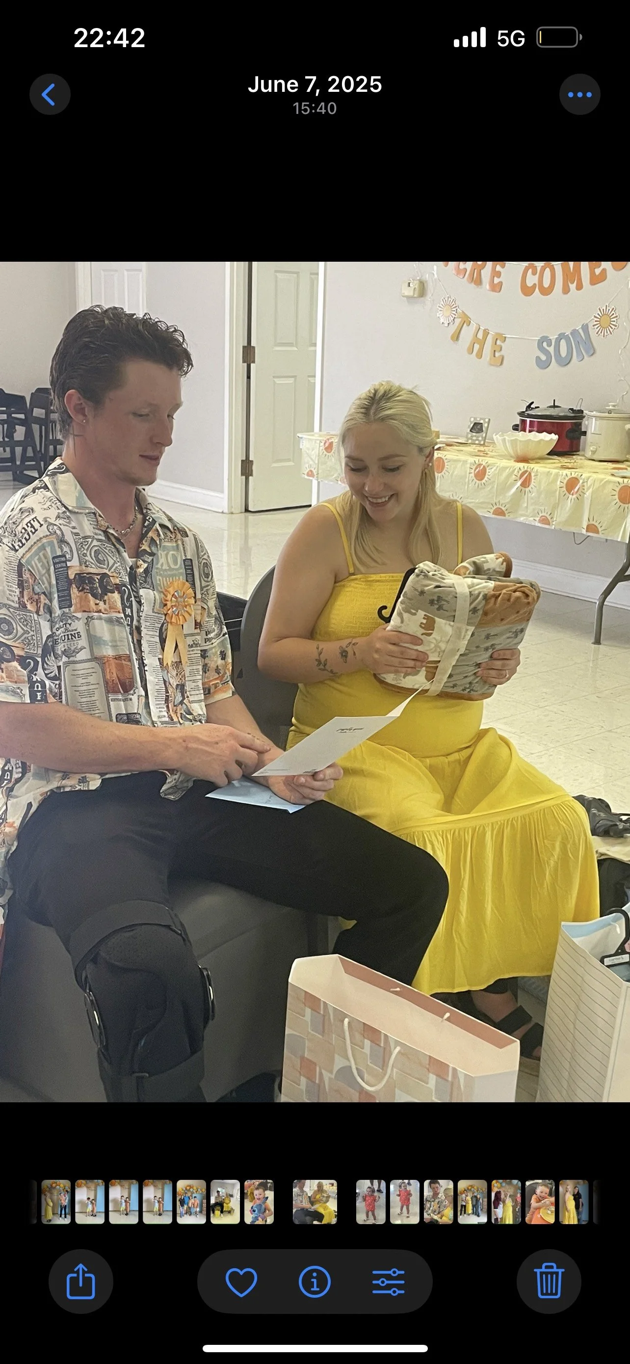 A man and a woman sit together at a celebration, with the woman holding a gift and a card. The man is looking at a piece of paper. The woman is smiling and dressed in a yellow dress. The background shows a decorated table with a banner, and gifts on the floor.