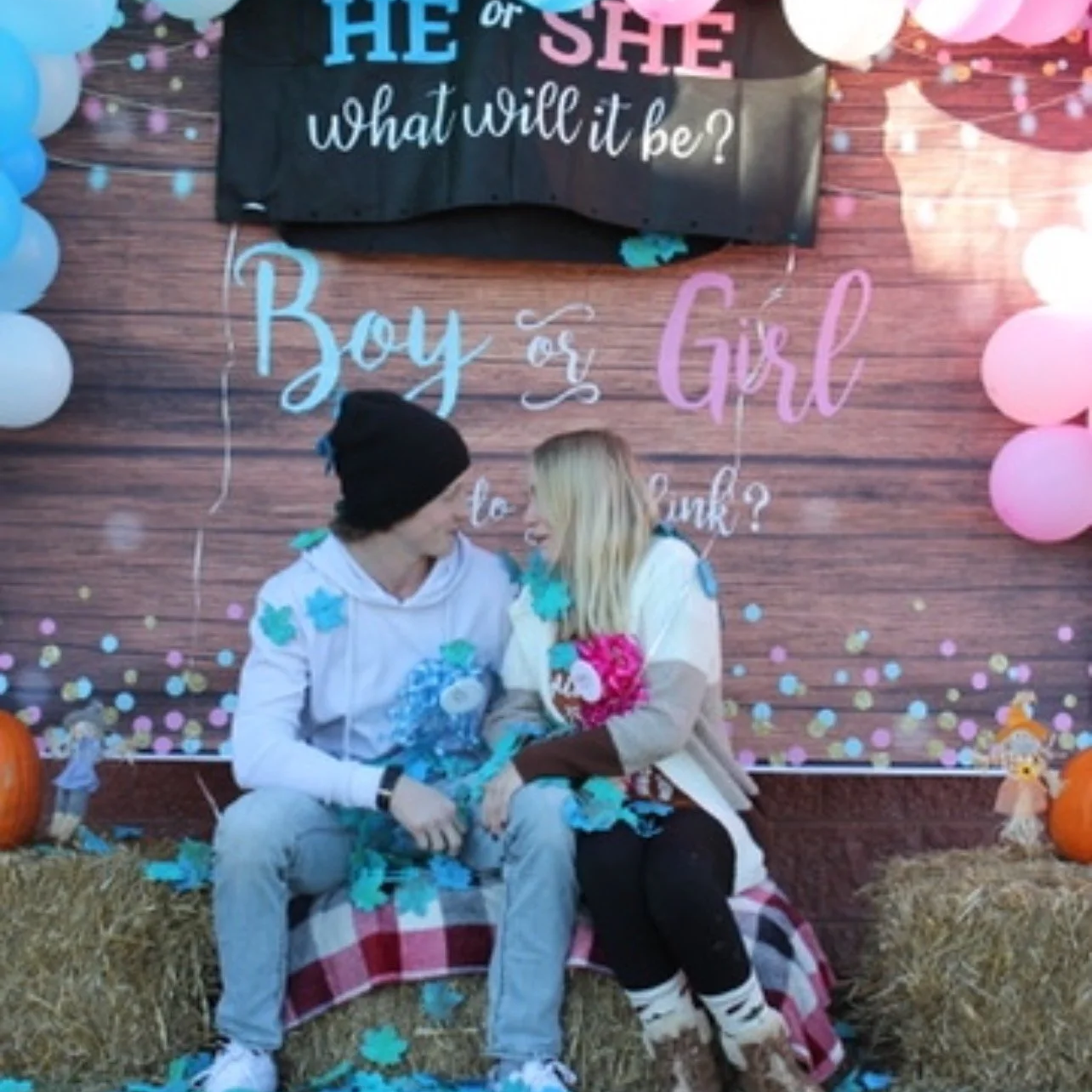 A couple sits on hay bales with fall pumpkins and decorations, at a gender reveal party with a backdrop that says, 'He or She, What will it be? Boy or Girl, do you think?' Pink and blue balloons and confetti surround them.