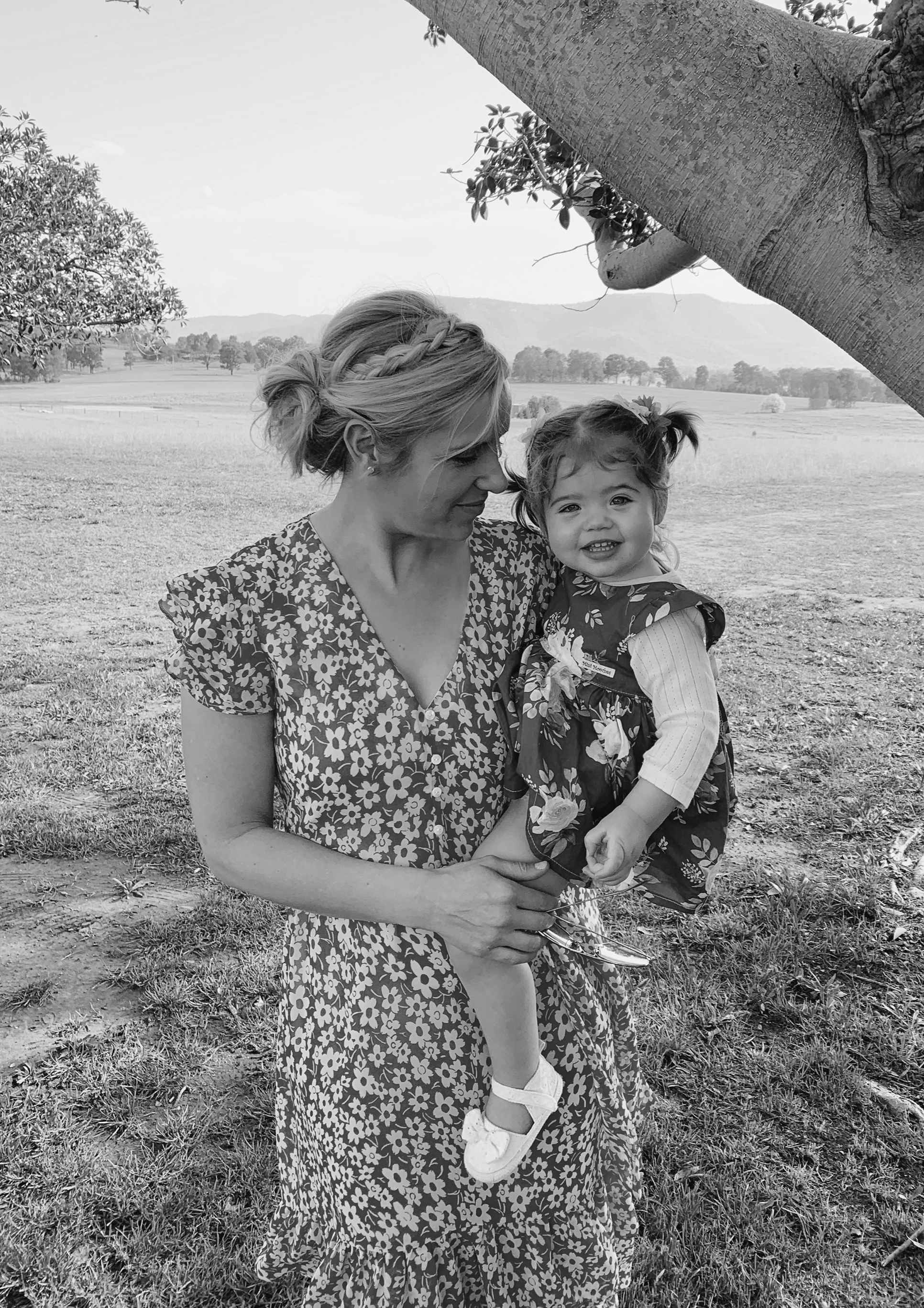 A woman with braided hair and a floral dress holding a young girl with pigtails and a dress, standing outdoors on grass with trees and hills in the background.