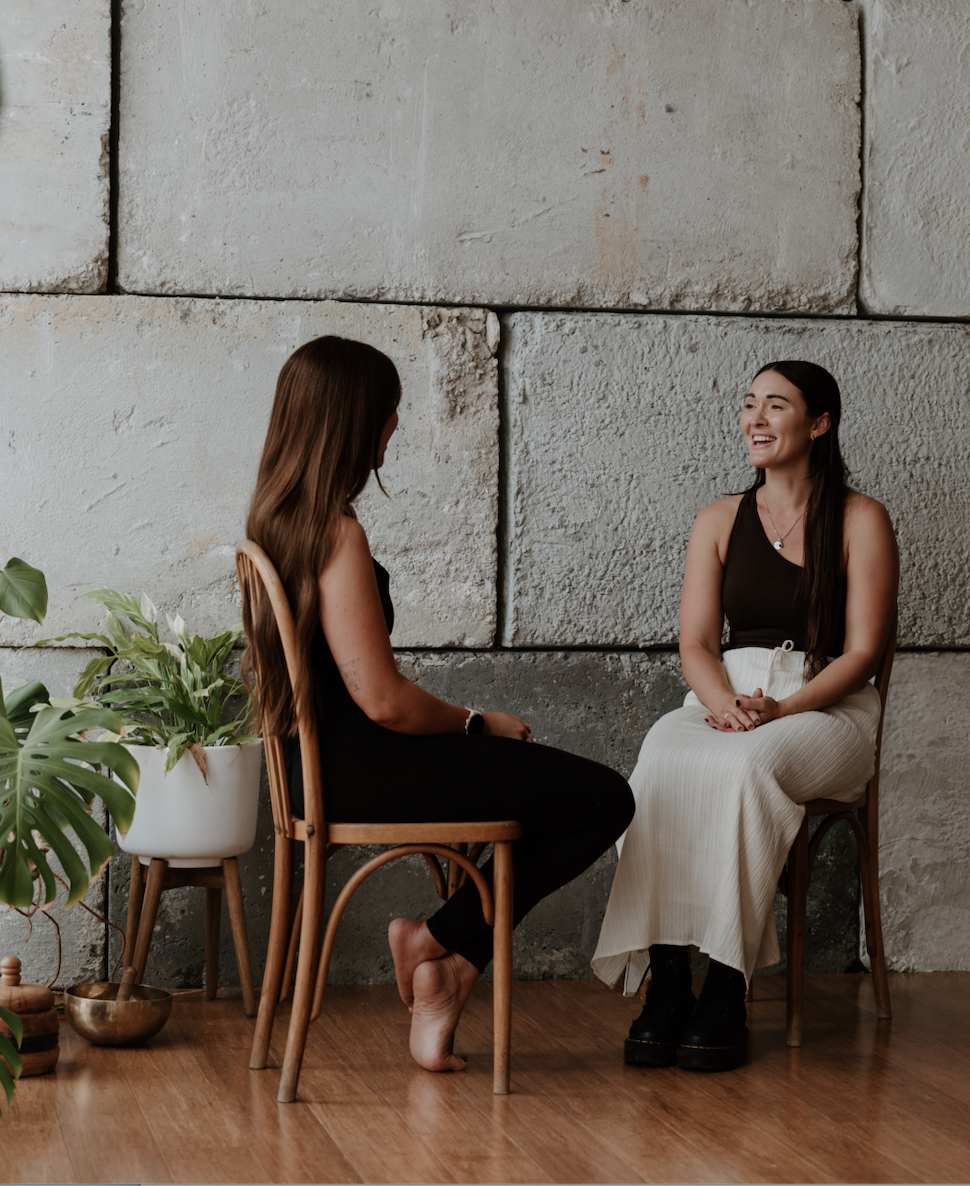 Two women having a conversation indoors, seated on wooden chairs against a concrete wall, with a large potted plant nearby.