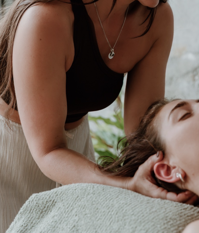 A woman giving a massage to another woman lying face-up, with closed eyes, on a massage table covered with a towel.