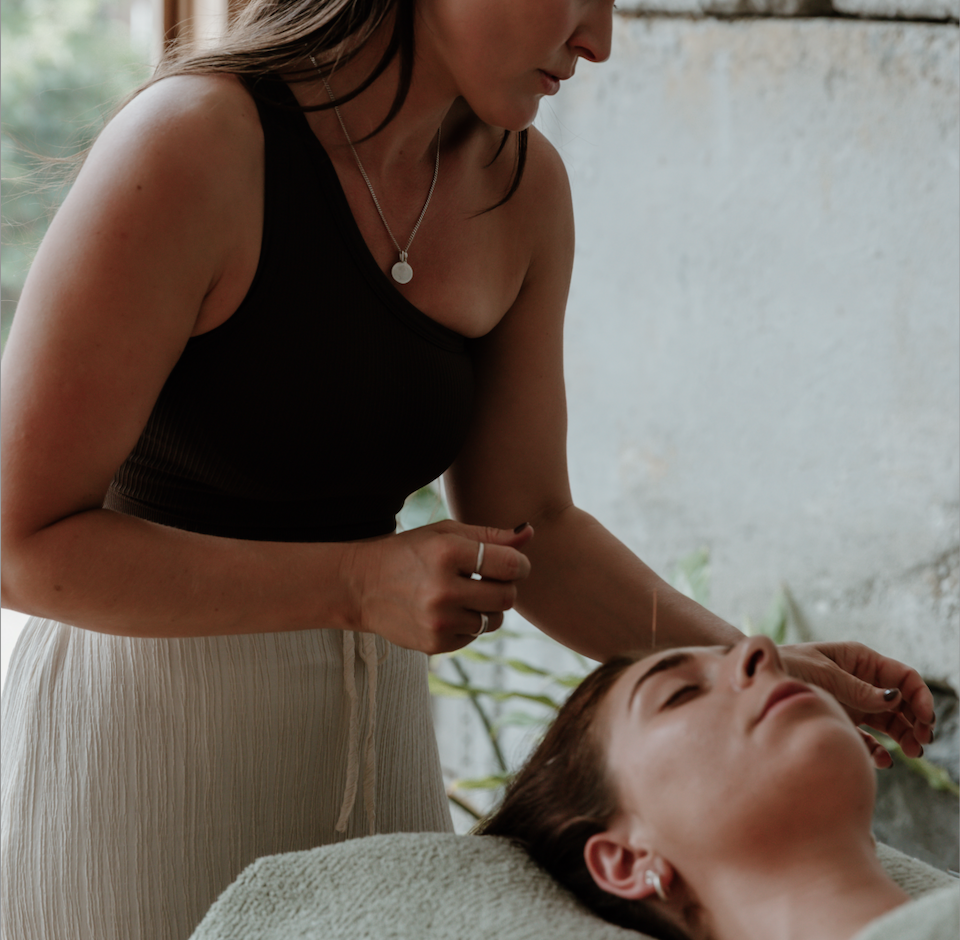 A woman giving acupuncture treatment to a person lying down on a bed, with the woman gently inserting a needle into the patient's arm.