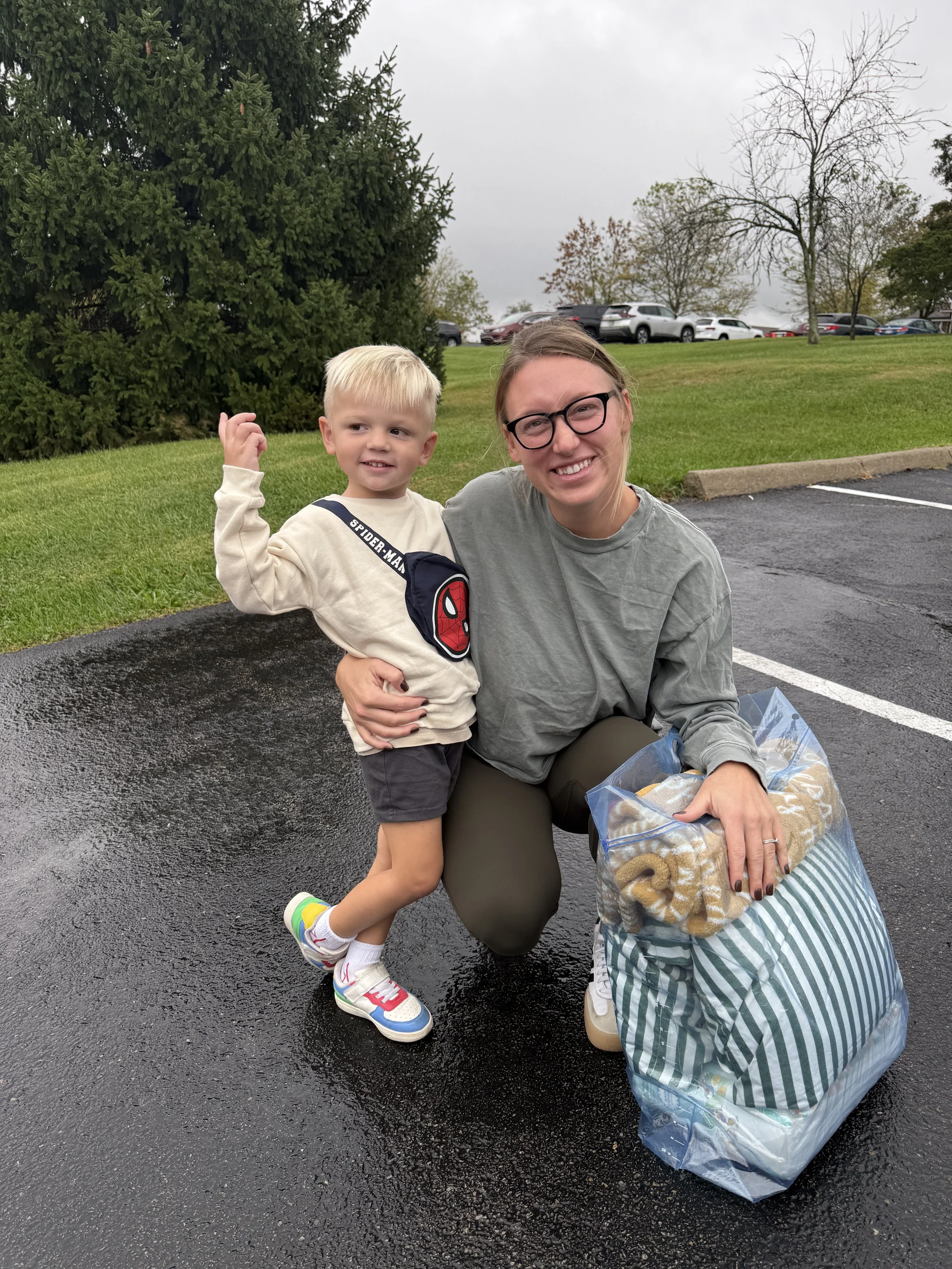 A woman with glasses smiling and a young boy with blond hair, wearing a Spider-Man sweatshirt, standing outdoors on a wet parking lot, with the woman holding a large bag and the boy making a fist in the air. Overcast sky with trees and parked cars in the background.