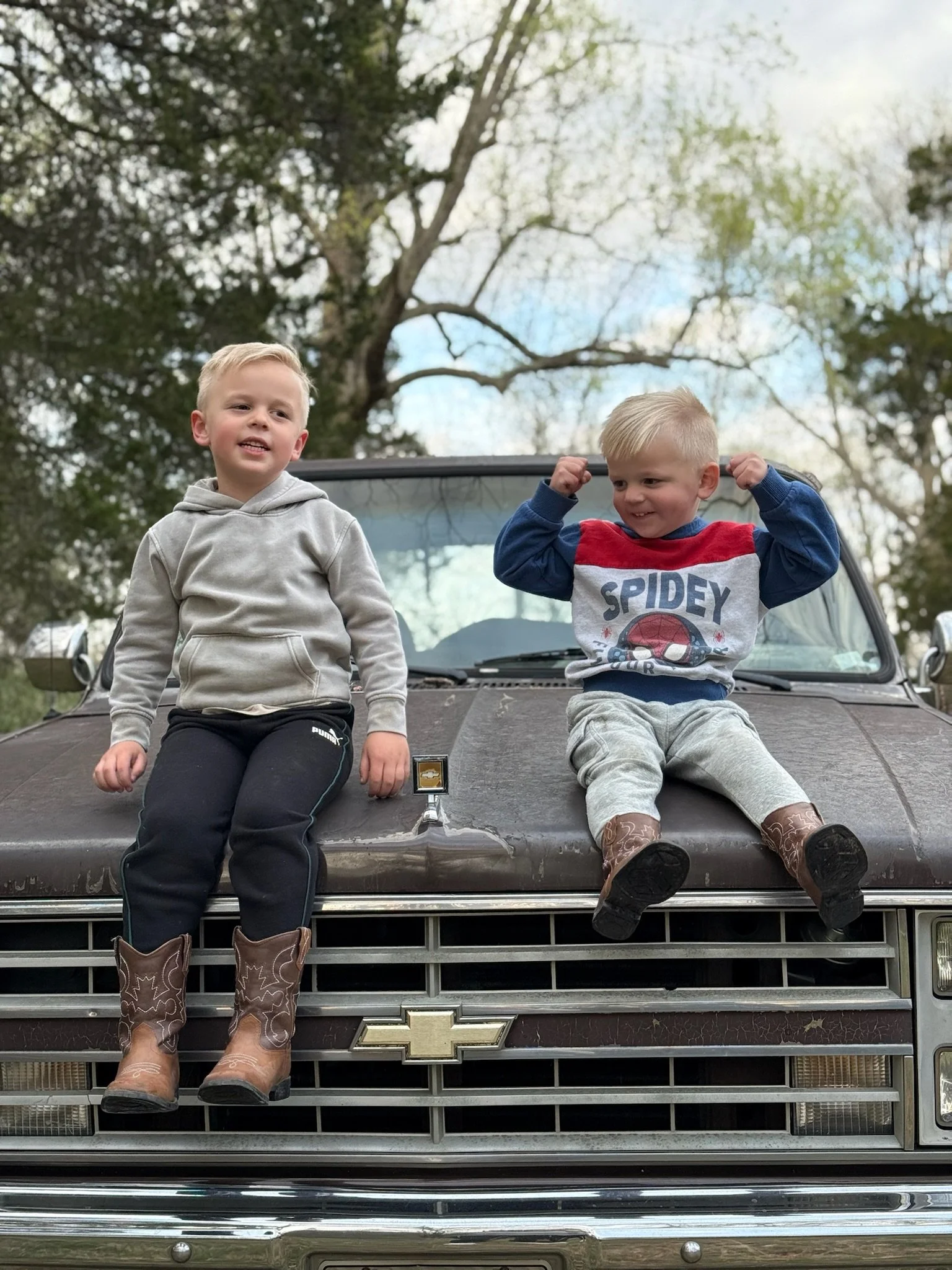 Two young boys sitting on the hood of a vintage Chevrolet vehicle outdoors, with trees and a partly cloudy sky in the background. One boy is wearing a gray hoodie and black pants, while the other is wearing a Spiderman sweater and light-colored pants. Both boys are wearing cowboy boots.