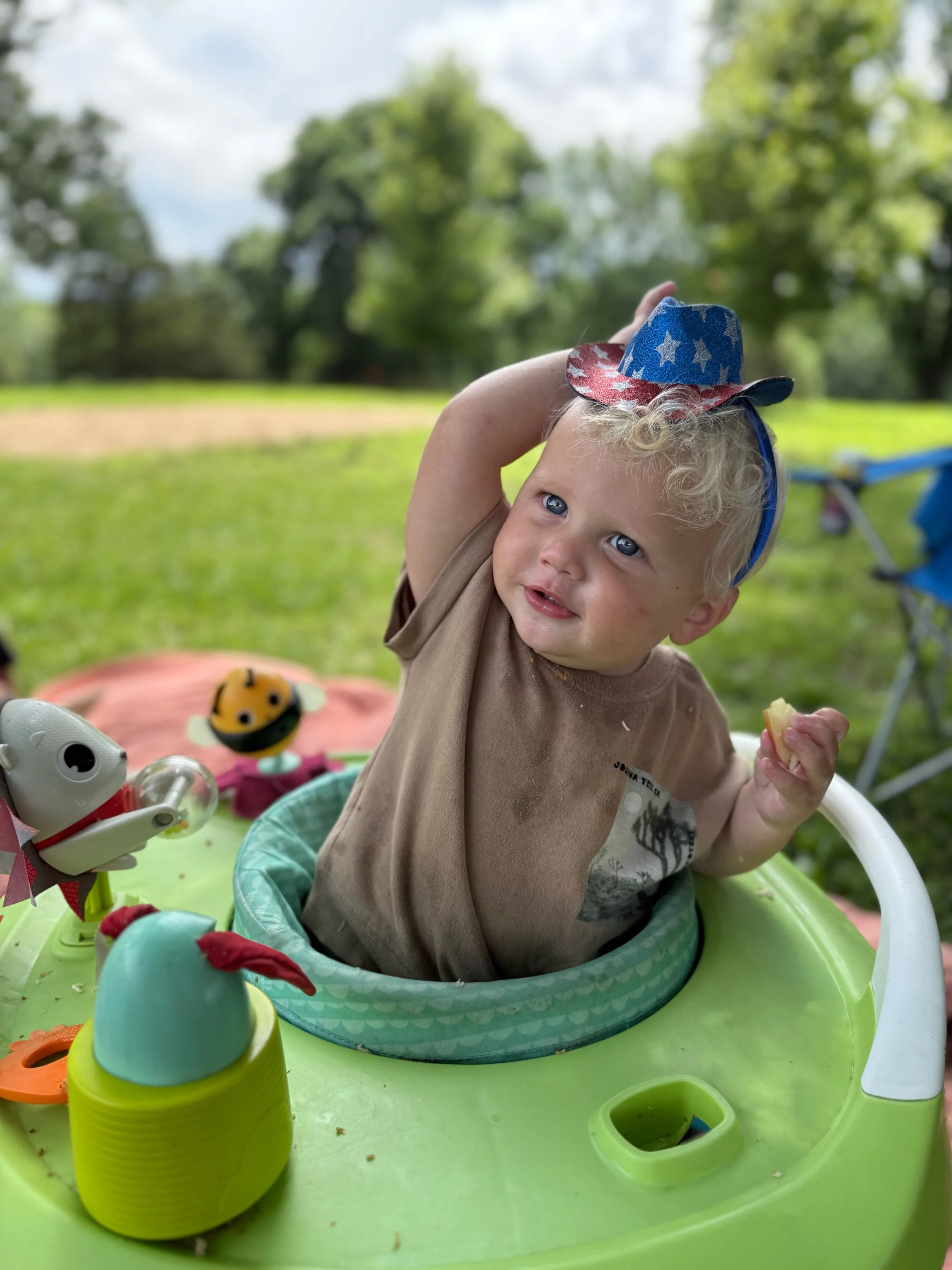 A young child with curly blonde hair and blue eyes wearing a brown shirt, sitting in a green and white baby walker outdoors. The child is holding a piece of food in one hand and adjusting a festive Uncle Sam hat on their head with the other. The background features green trees and a partly cloudy sky, and the walker is decorated with toys and colorful items.