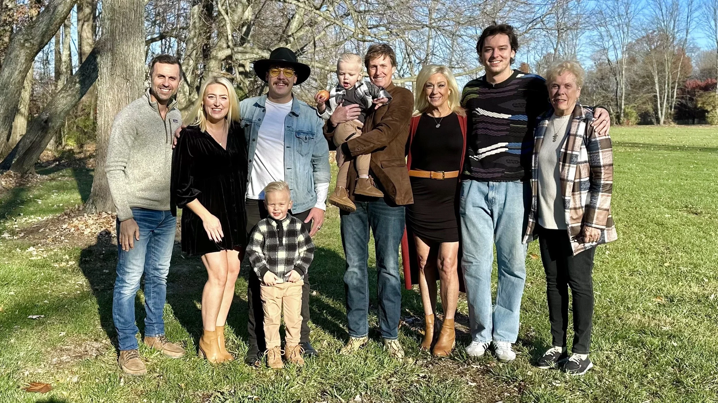 A group of ten people, including children, standing outdoors on grass in front of trees on a sunny day. They are smiling and dressed casually, with some wearing jackets and boots.
