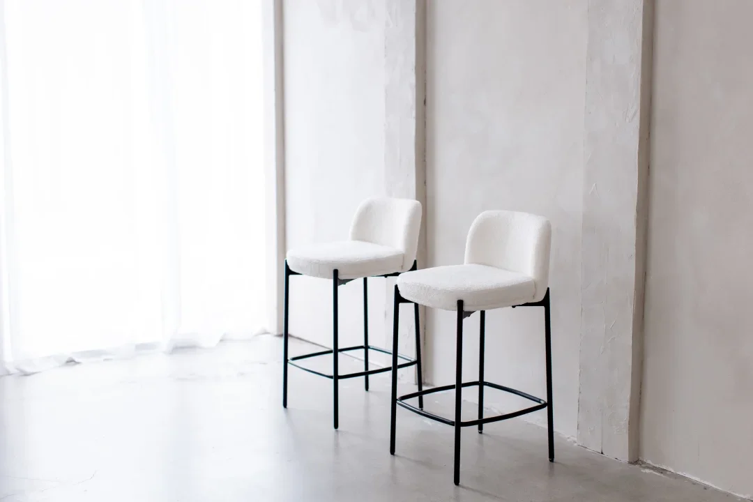 Two white upholstered chairs with black metal legs in a bright room with sheer white curtains and a light-colored wall.