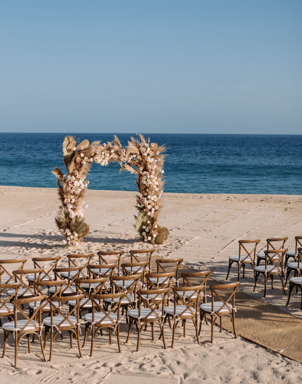 Empty chairs on a sandy beach facing a floral wedding arch with the ocean in the background.