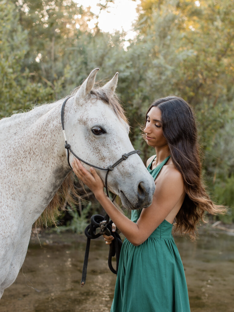 A woman in a green dress gently touching the face of a white horse by a river in Orange County, with trees and sunlight in the background.