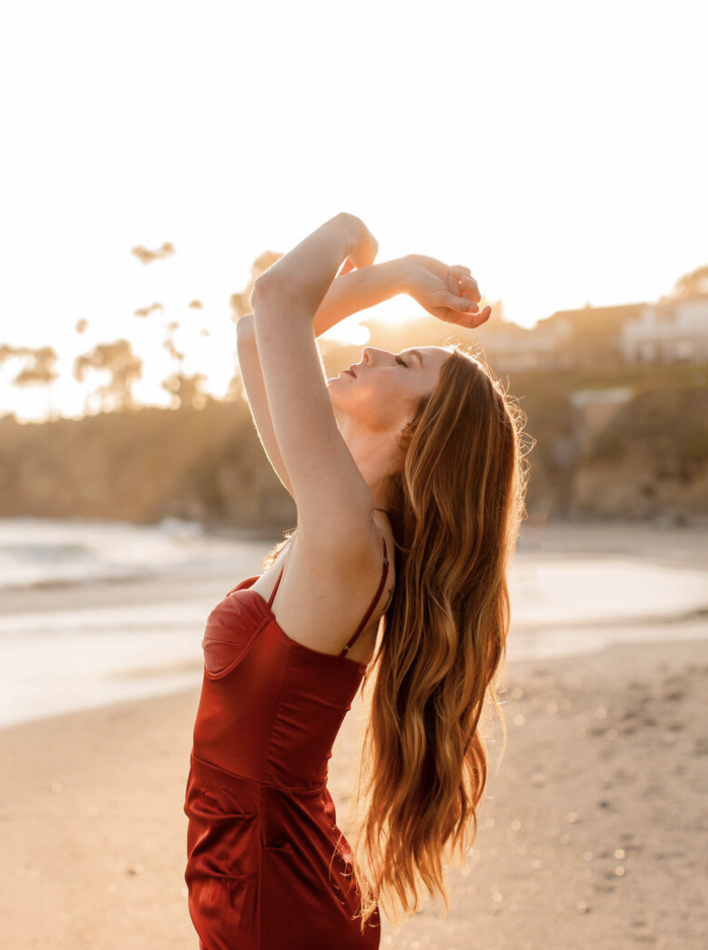 A woman with long red hair in a red dress in Laguna beach at sunset, striking a pose with arms raised above her head and eyes closed.