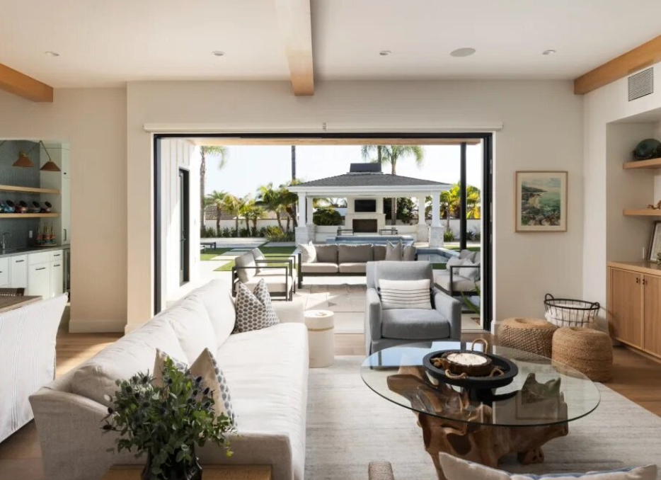 Living room with a large sliding glass door opening to an outdoor patio with seating and a gazebo, decorated with palm trees and a lawn.