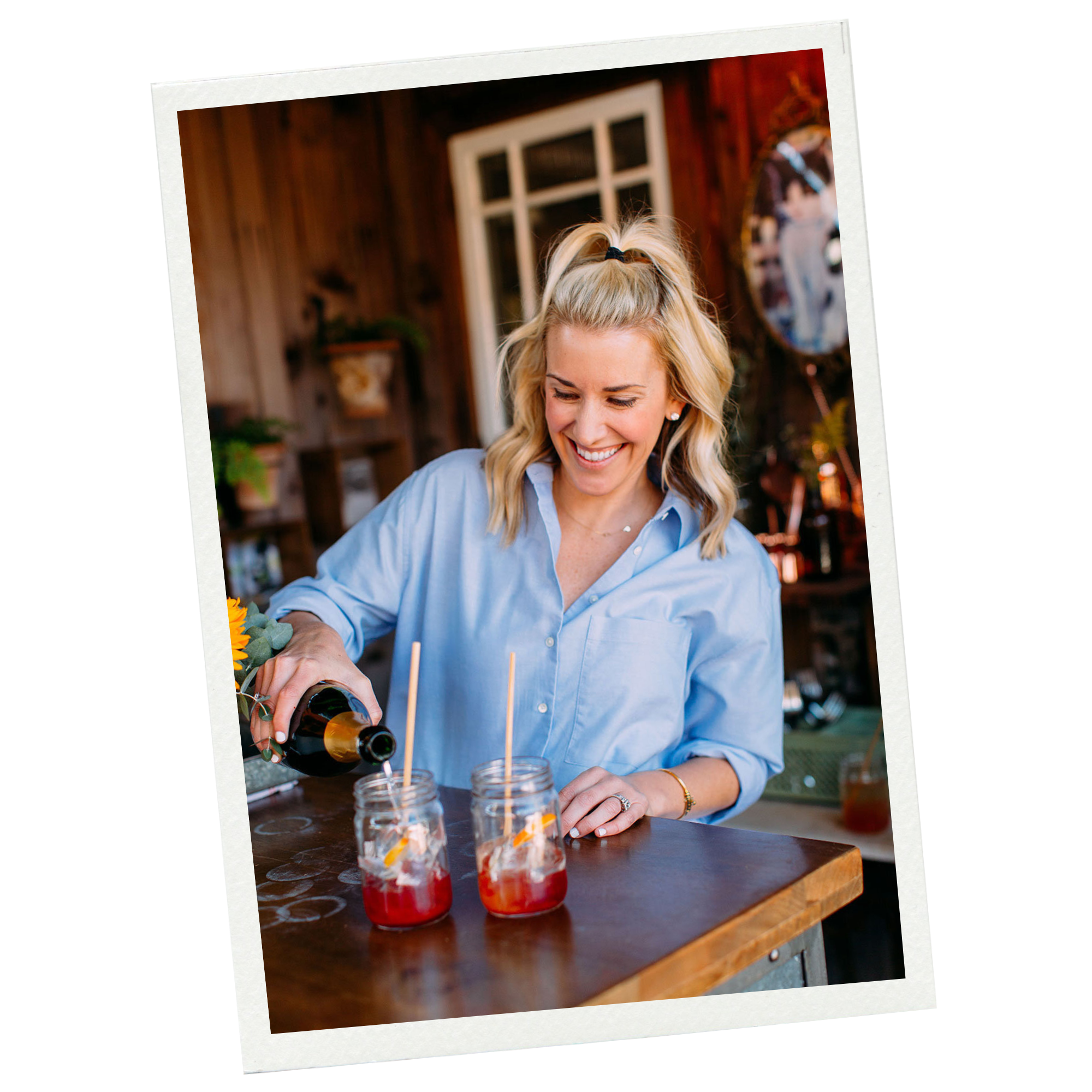 A woman with blonde hair in a half-up ponytail, smiling, wearing a light blue button-down shirt, pouring a drink into mason jars with straws and fruit slices on a wooden table in a cozy, rustic setting.