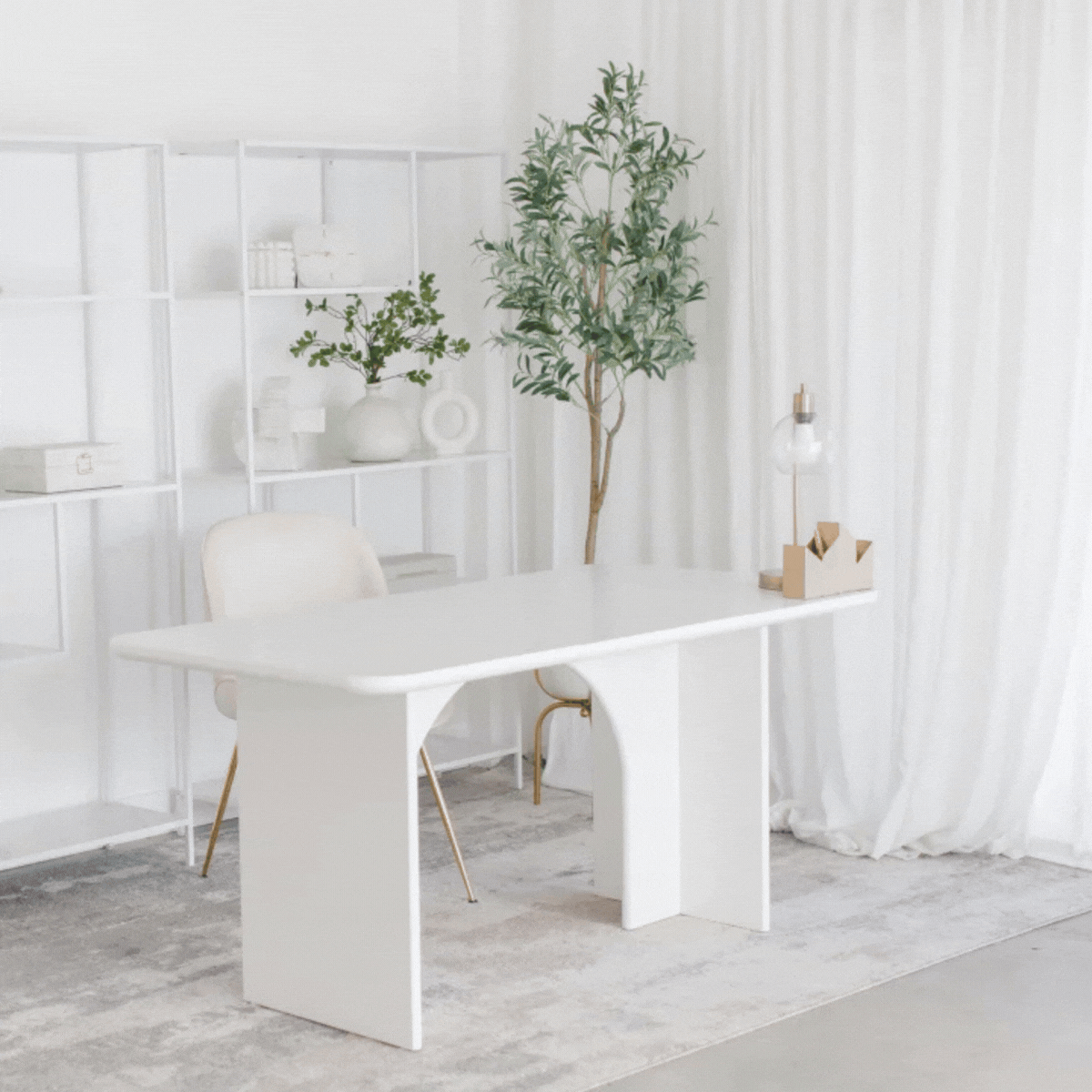 Minimalist office space with white desk, white chair, large green potted plant, white bookshelf with decor, and white curtains.