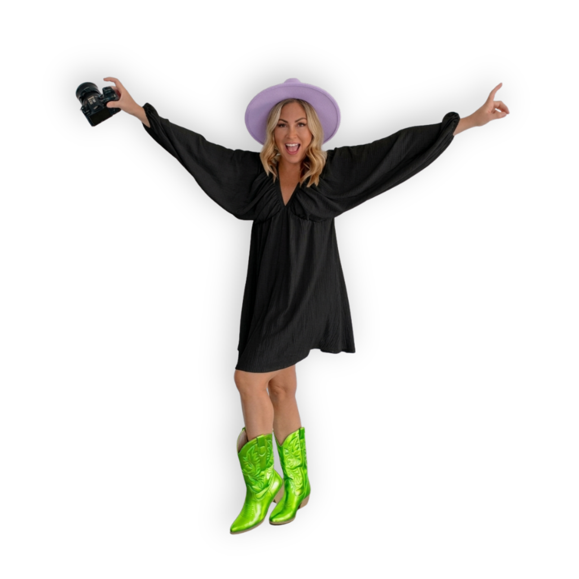 Woman dressed in a black dress, wearing bright green cowboy boots, a lavender wide-brim hat, and holding a camera, posing against a plain black background with arms outstretched and smiling.