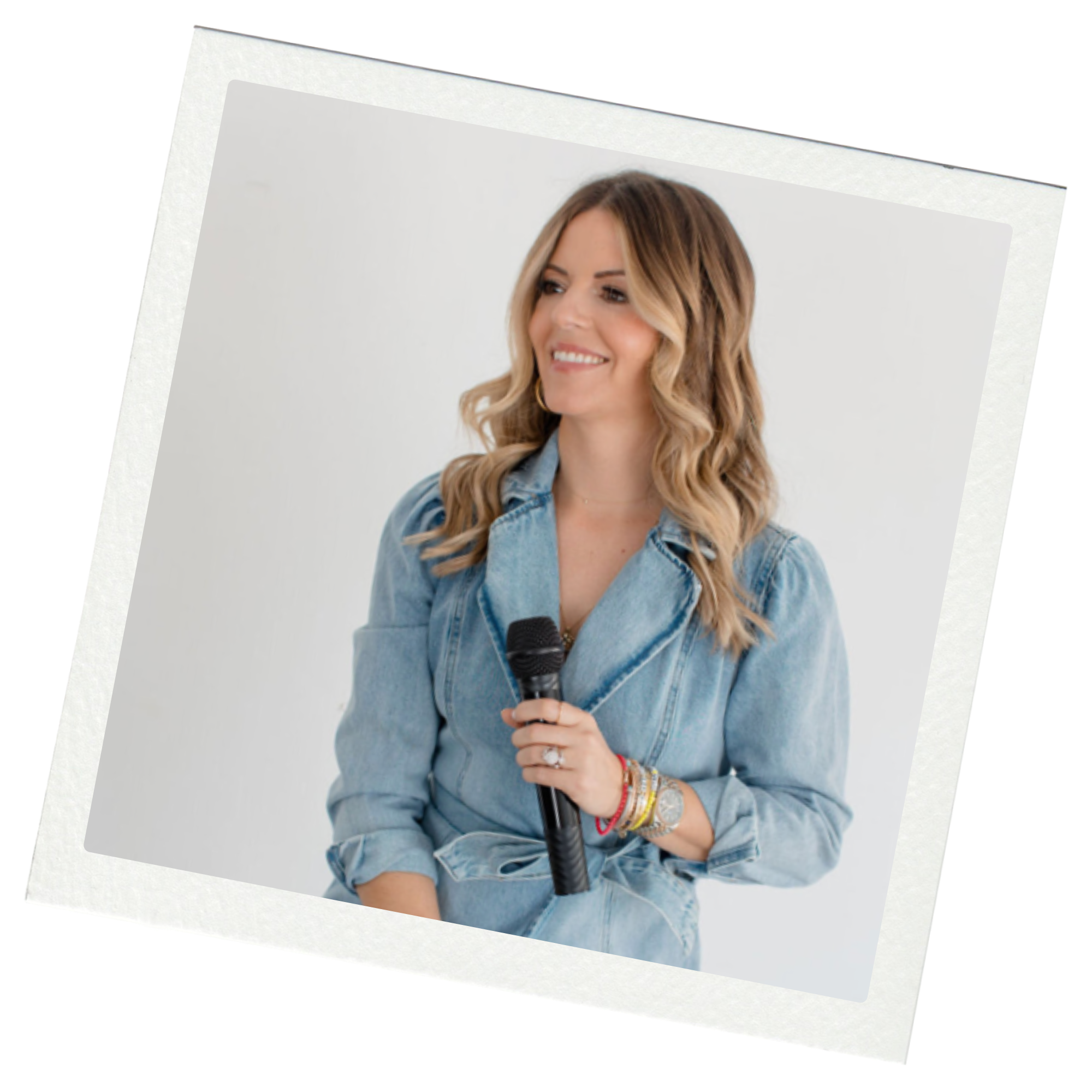 A smiling woman with wavy, shoulder-length hair, wearing a denim jacket, holding a microphone, sitting against a plain white background.