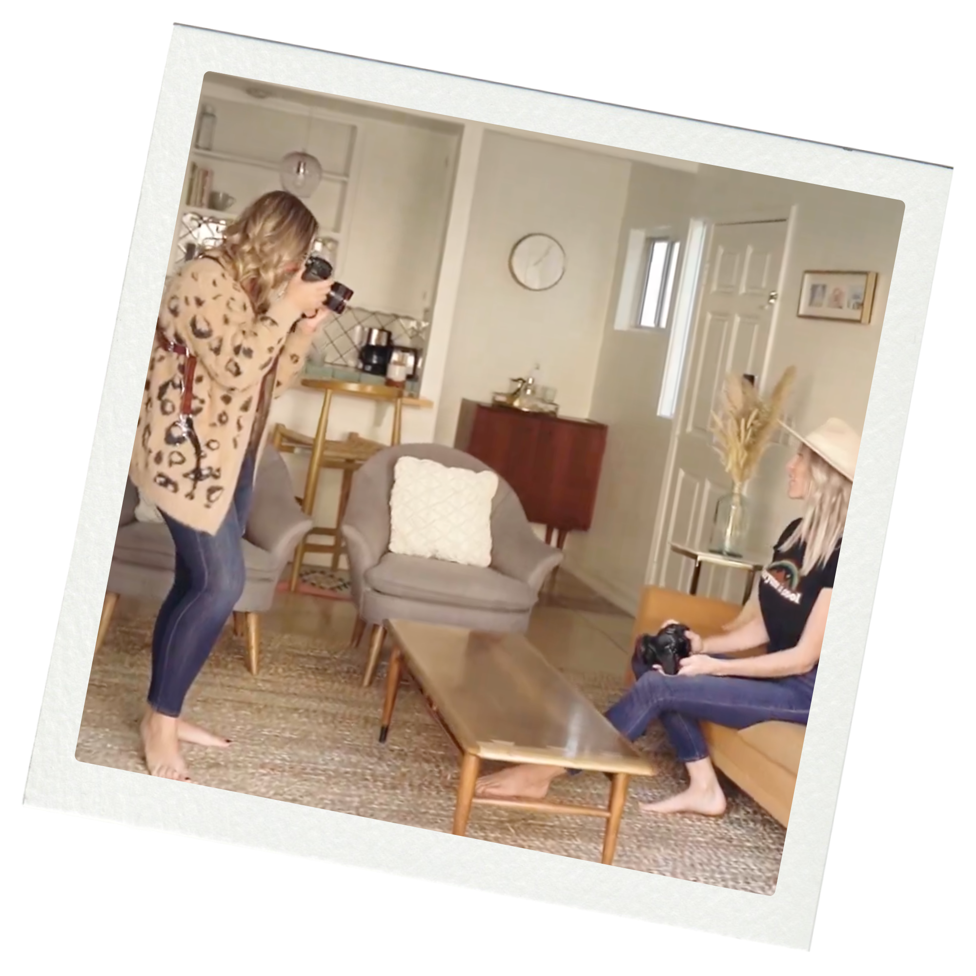 Two women with cameras in a living room, one standing, one sitting on a sofa, preparing for a photo shoot.
