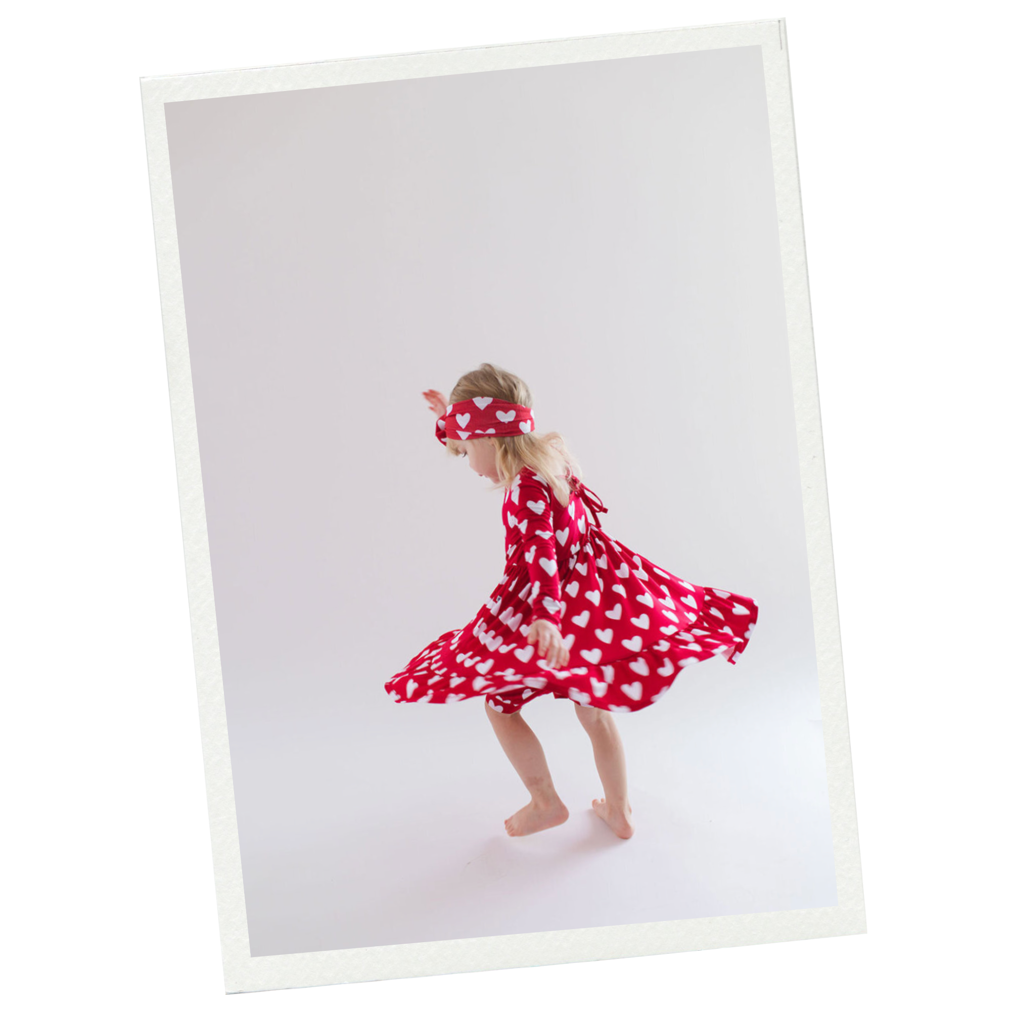 Young girl in a red dress with white hearts, spinning and twirling barefoot against a plain white background, wearing a matching headband.