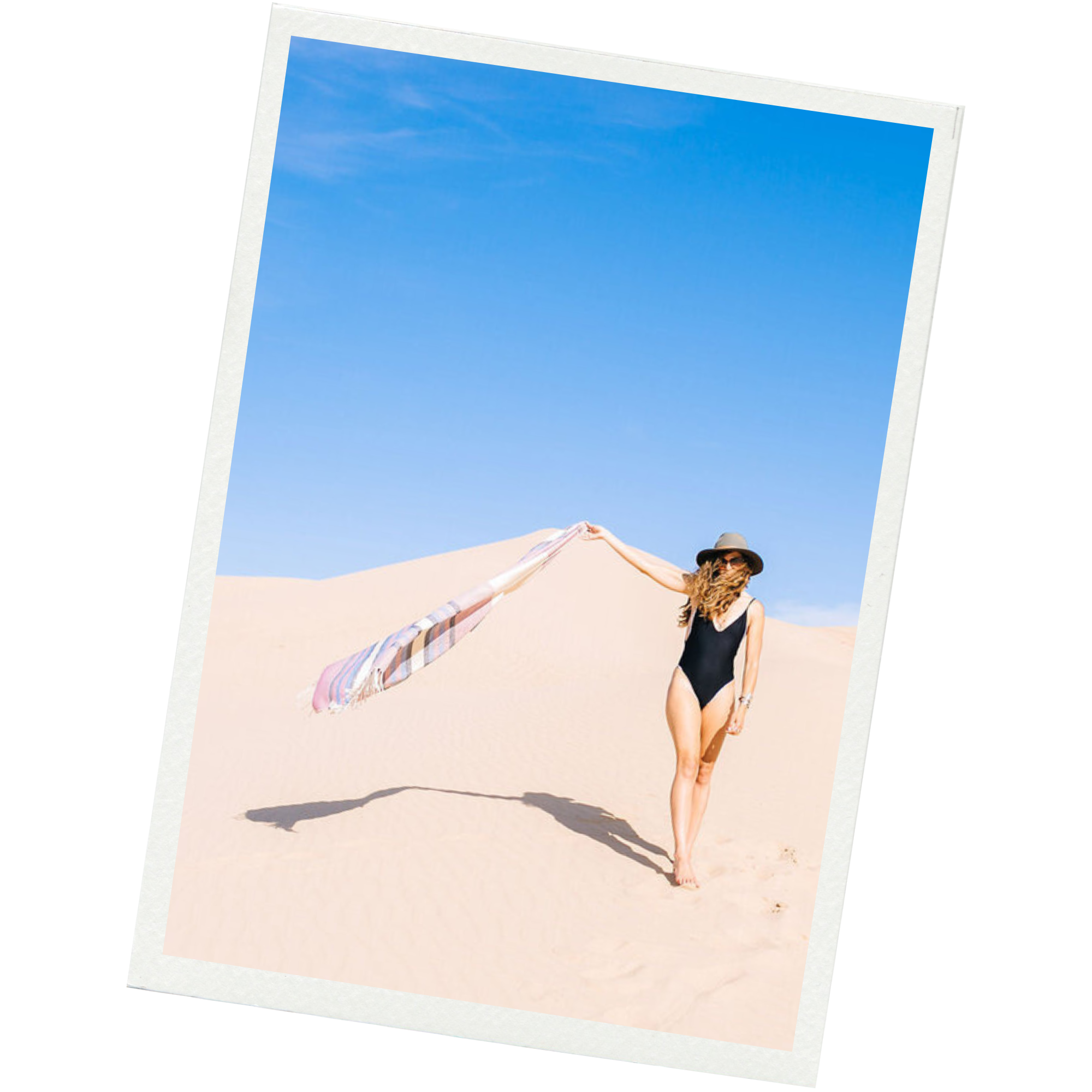 Woman in black swimsuit and wide-brimmed hat holding a striped towel in a sandy desert with clear blue sky.