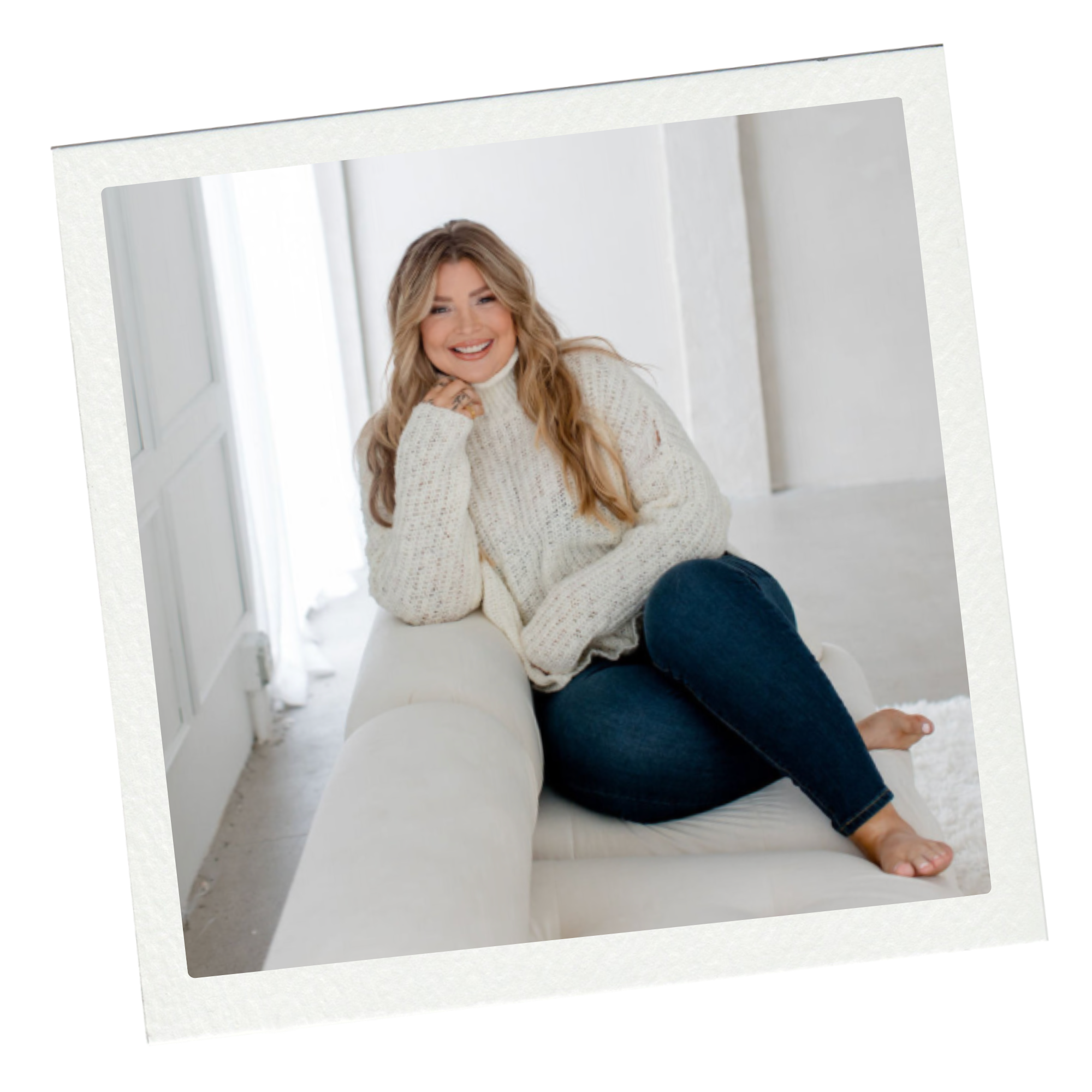 A woman with long wavy blonde hair sitting comfortably on a white sofa, smiling, in a bright room with white walls and a window with sheer curtains.
