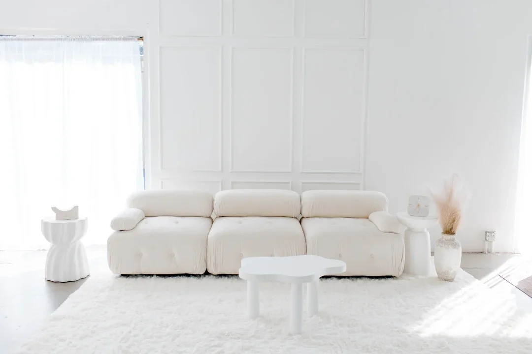 Bright living room with all-white decor, including a large white sofa, a white coffee table, two white side tables, and a vase with dried pampas grass.