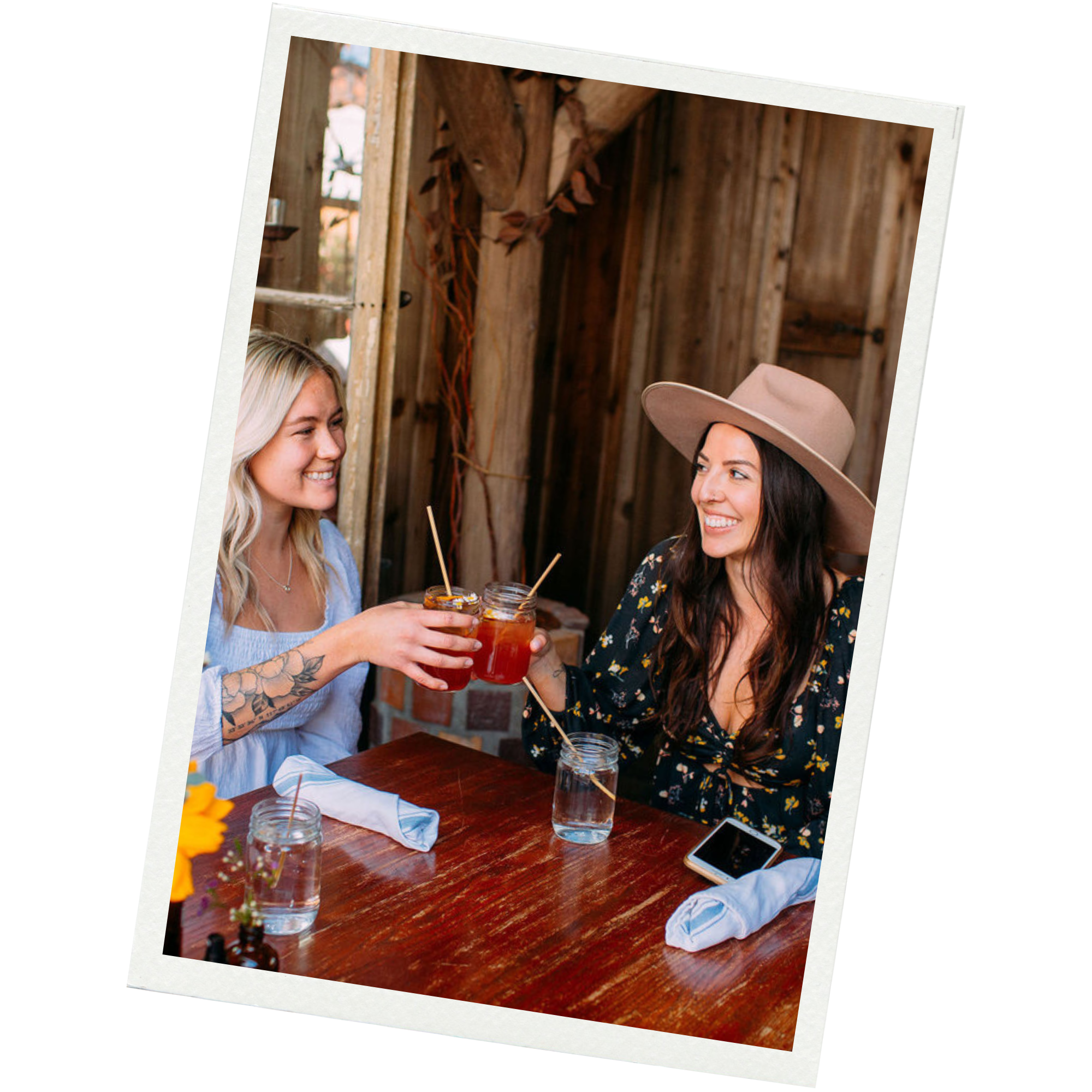 Two women smiling and enjoying drinks at a wooden table in a rustic setting, clinking mason jar glasses with drinks, one wearing a wide-brim hat.