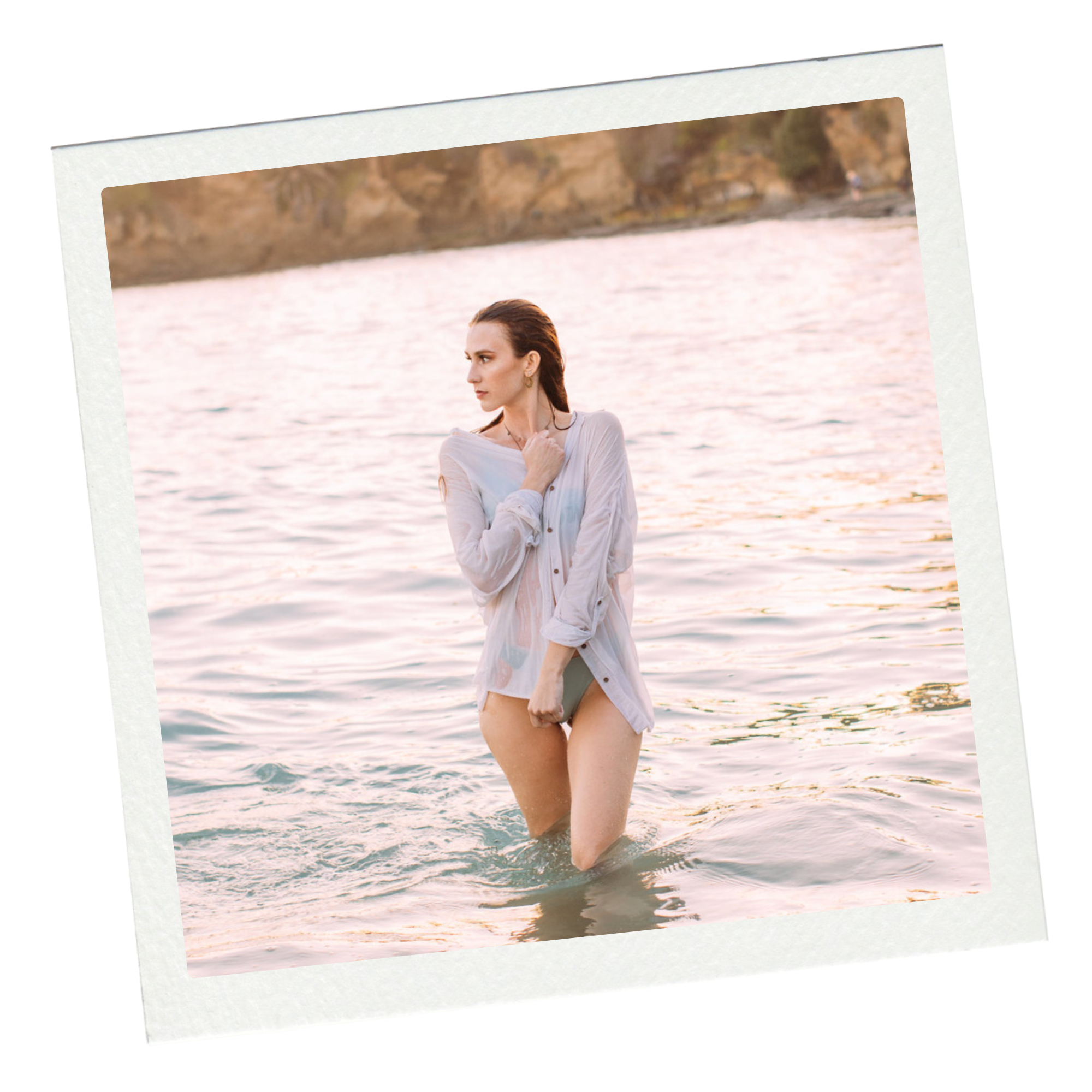A woman standing in the water at sunset, wearing a wet shirt and holding a phone.