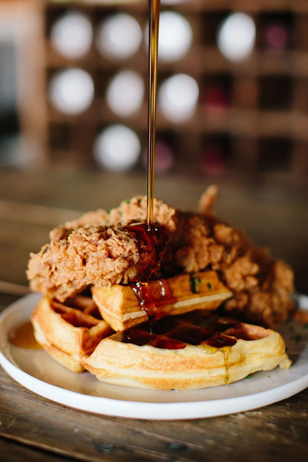 A plate of waffles topped with fried chicken, syrup being poured over it, with a blurred background of a wine rack.