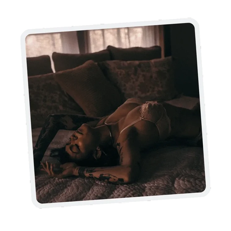 A woman with tattoos lying on a bed with pillows, wearing lace lingerie, in a dimly lit room.