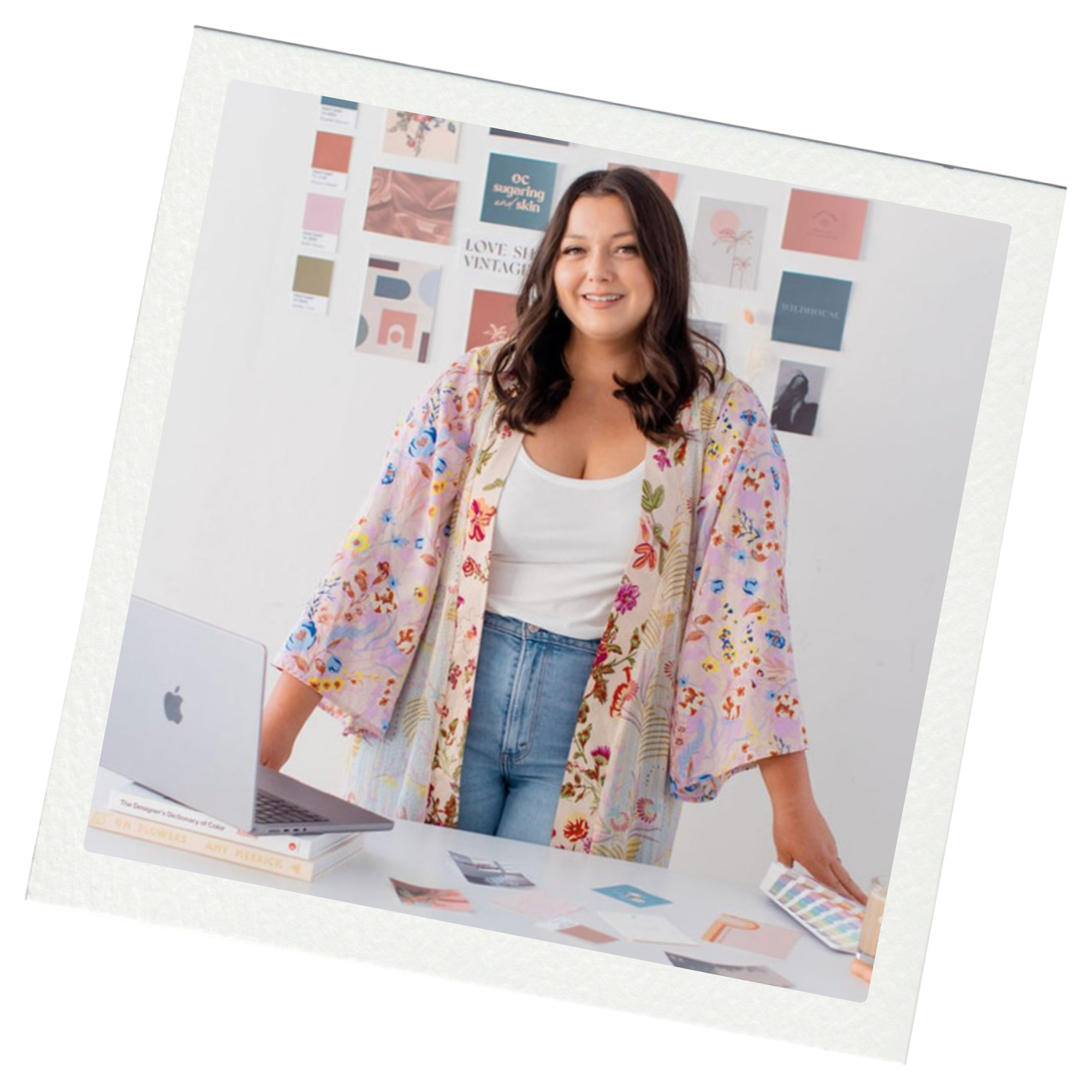 A woman with shoulder-length brown hair, wearing a white top and a colorful floral kimono, standing behind a creative workspace with design materials, a laptop, and wall art in the background.