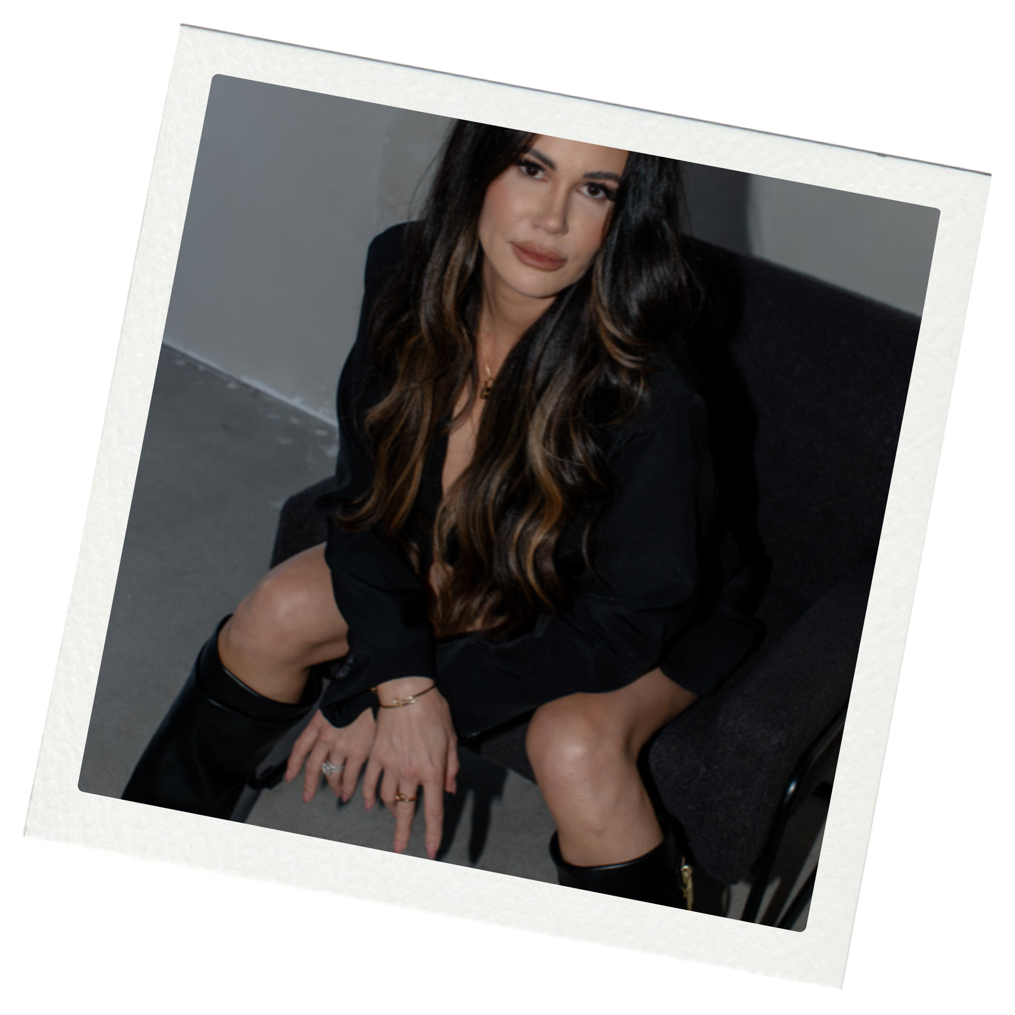 A woman with long dark wavy hair sitting on a black chair, wearing a black outfit and black boots, looking at the camera.