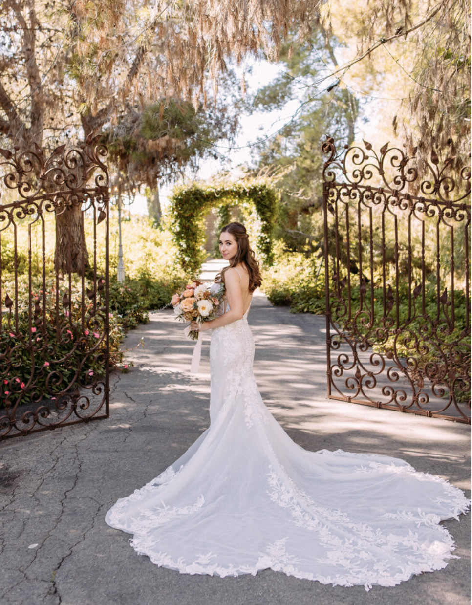 A bride in a white wedding gown holding a bouquet of flowers standing on a cracked asphalt pathway at a garden entrance, decorated with wrought iron gates. The pathway leads to a lush, sunlit garden with trees and a green archway in the background.