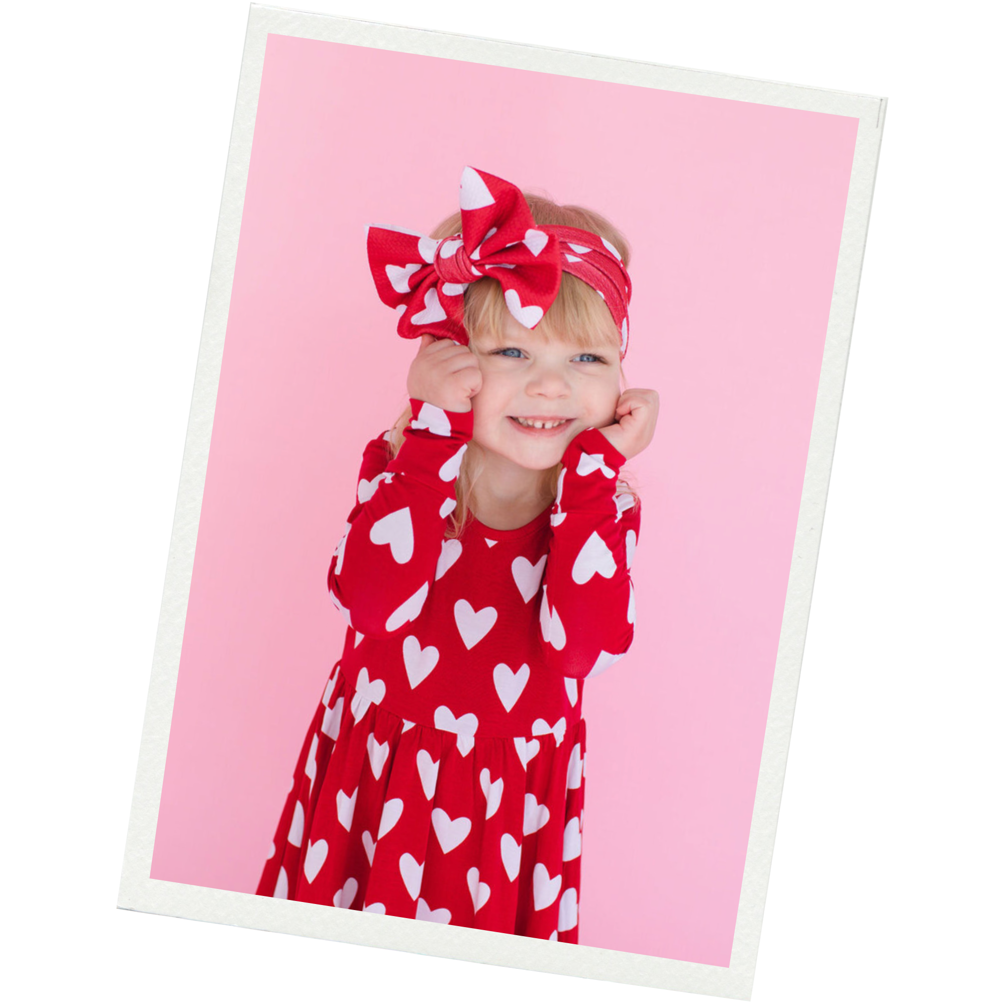 A young girl dressed in a red dress with white heart patterns, wearing a matching headband and wristbands, smiling while touching her cheeks against a pink background.