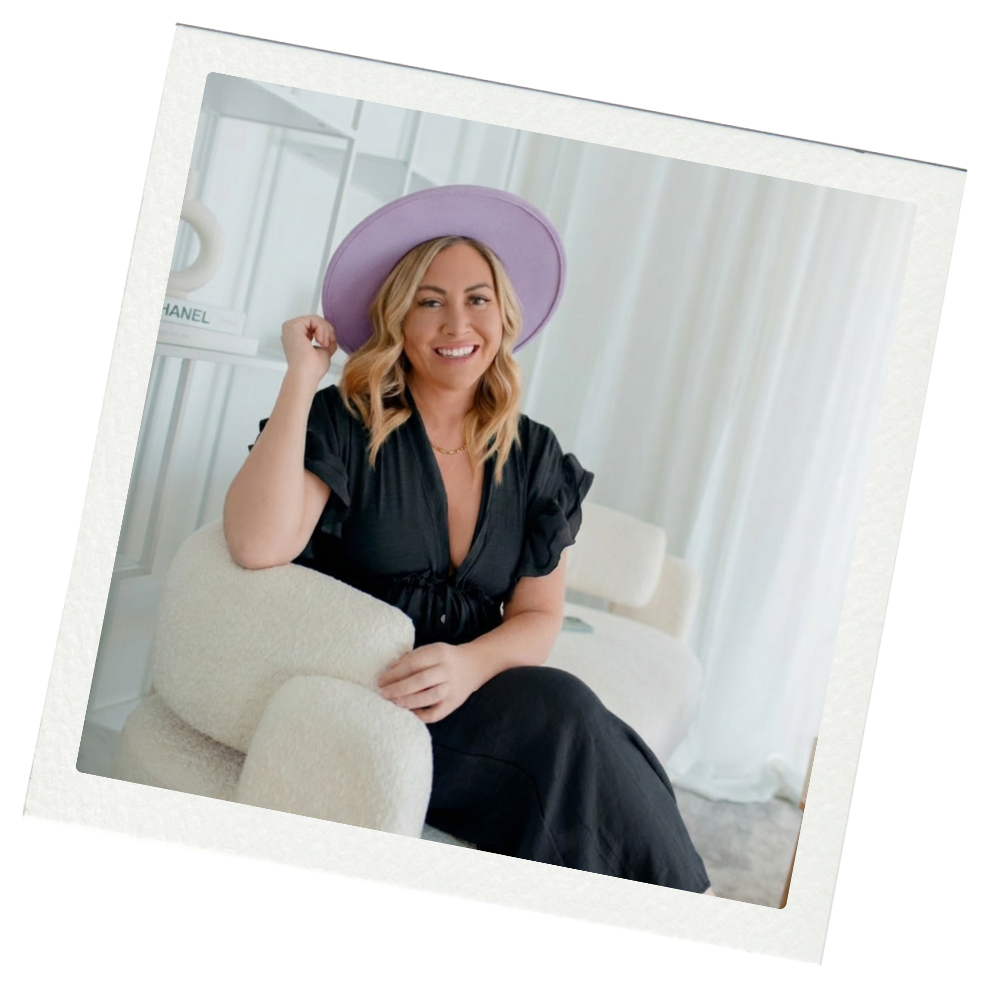 A woman with blonde hair, wearing a purple hat and a black dress, smiling while sitting on a cream-colored armchair in a bright, airy room with white curtains and shelves.