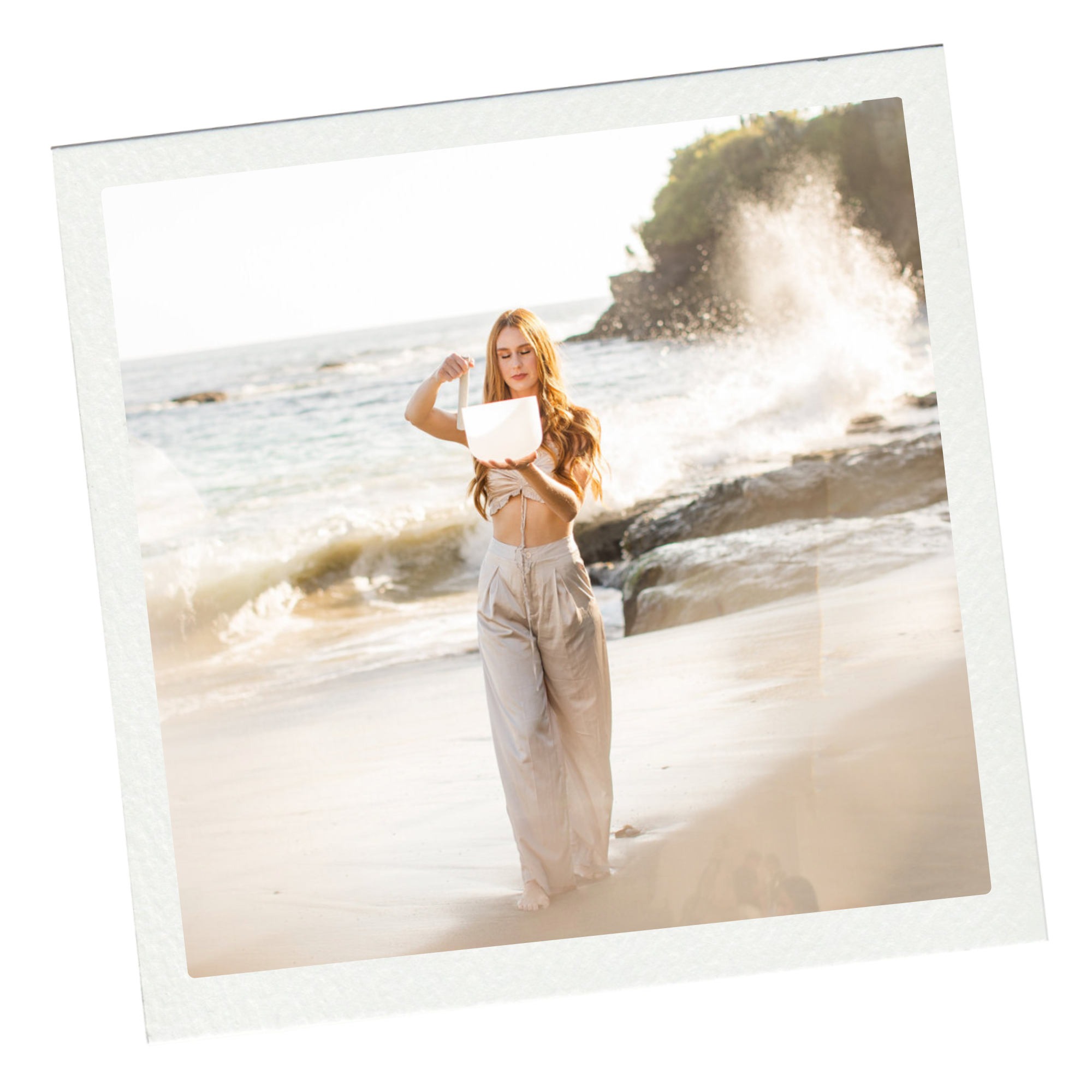 A woman with long red hair wearing beige wide-leg pants and a beige crop top standing on a beach, holding a white bucket, with ocean waves crashing against rocks in the background.