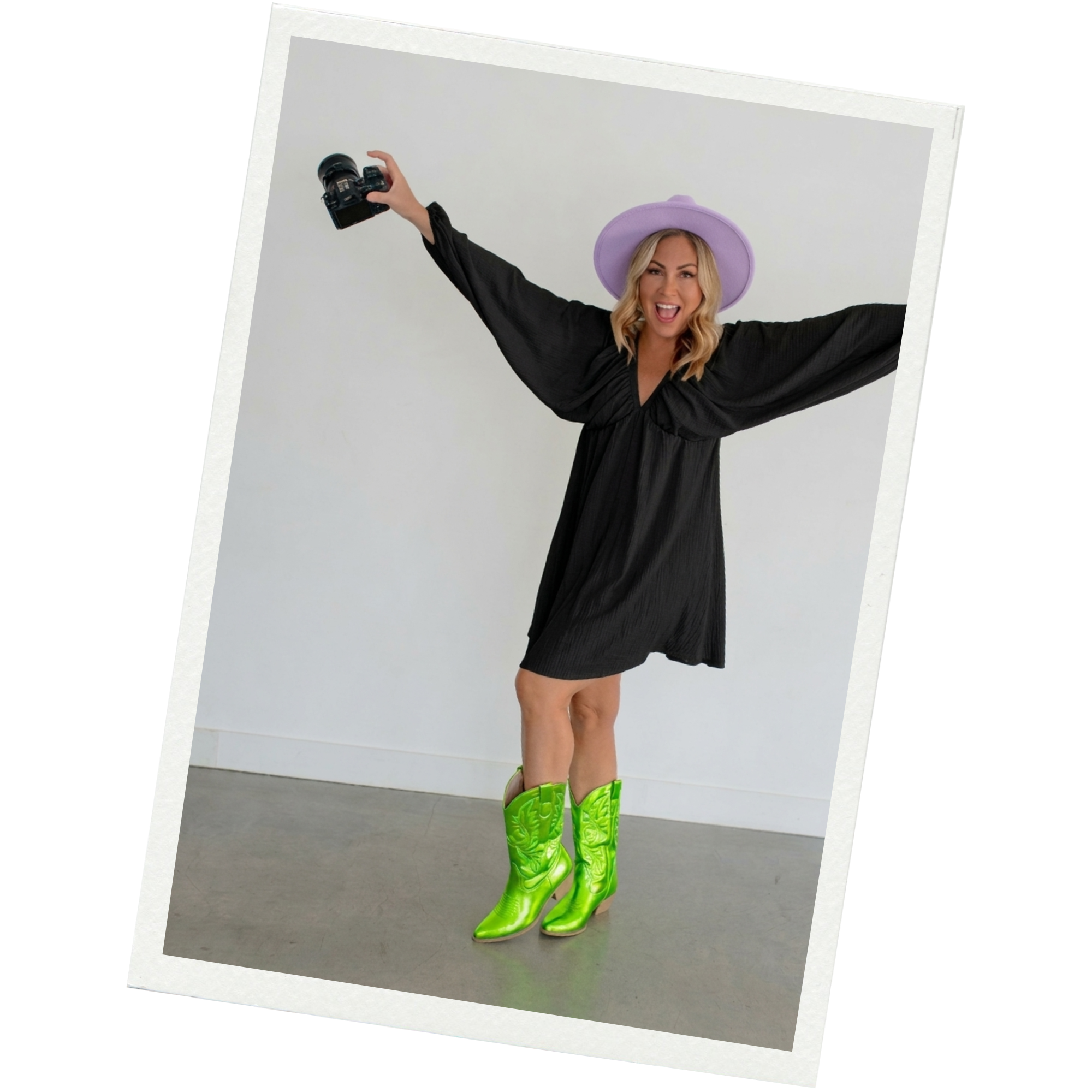 A woman in a black dress and neon green cowboy boots, wearing a lavender hat, is holding a camera in one hand and smiling with her arms outstretched against a plain white background.