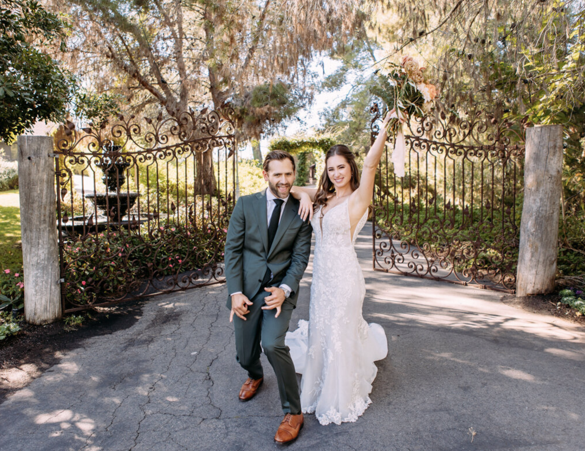A joyful bride and groom celebrating outdoors in a garden, with the bride holding a bouquet and the groom smiling, standing in front of a decorative iron gate.