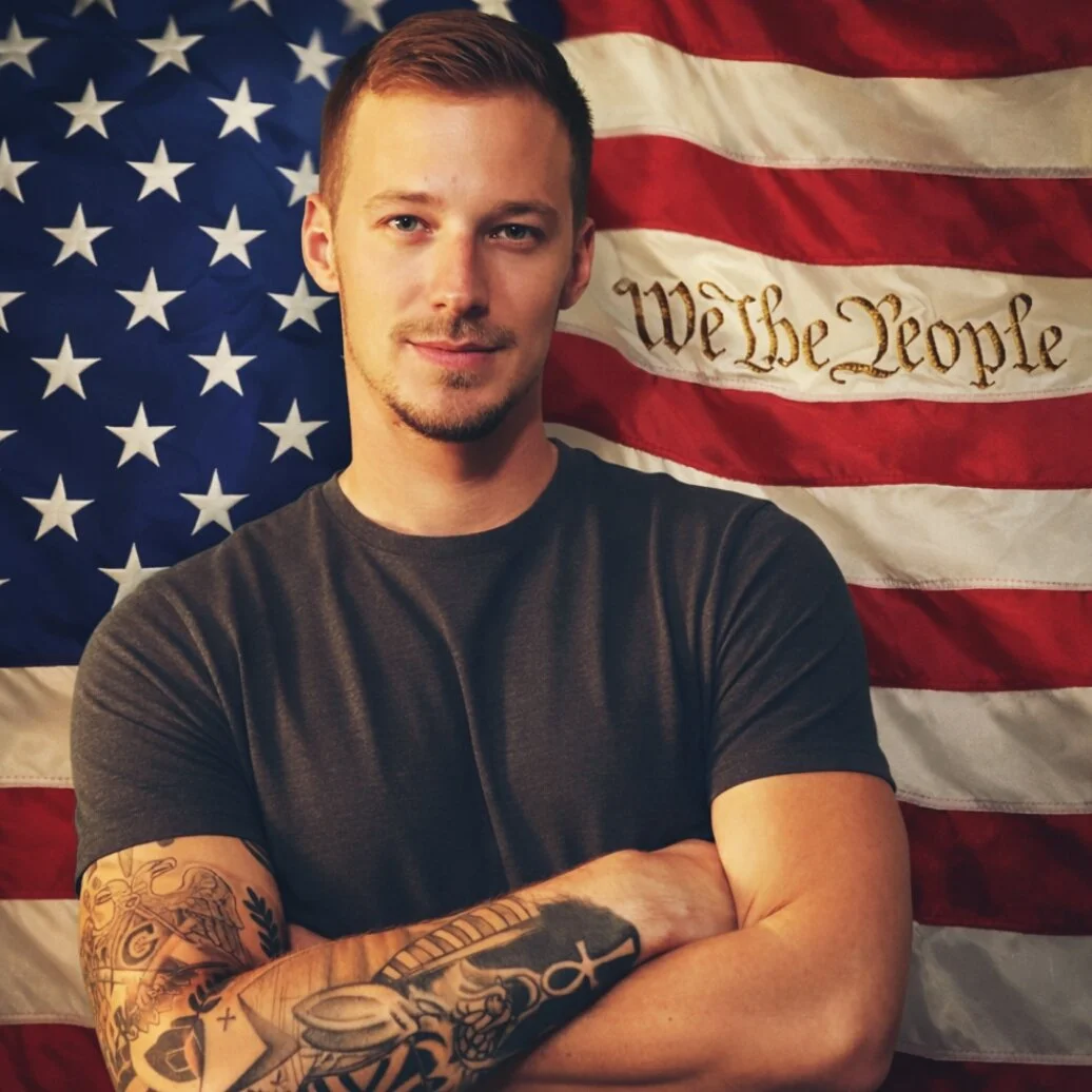 A young man with short hair and tattoos on his arms, standing with crossed arms in front of an American flag background. The flag has the words We the People embroidered on it.