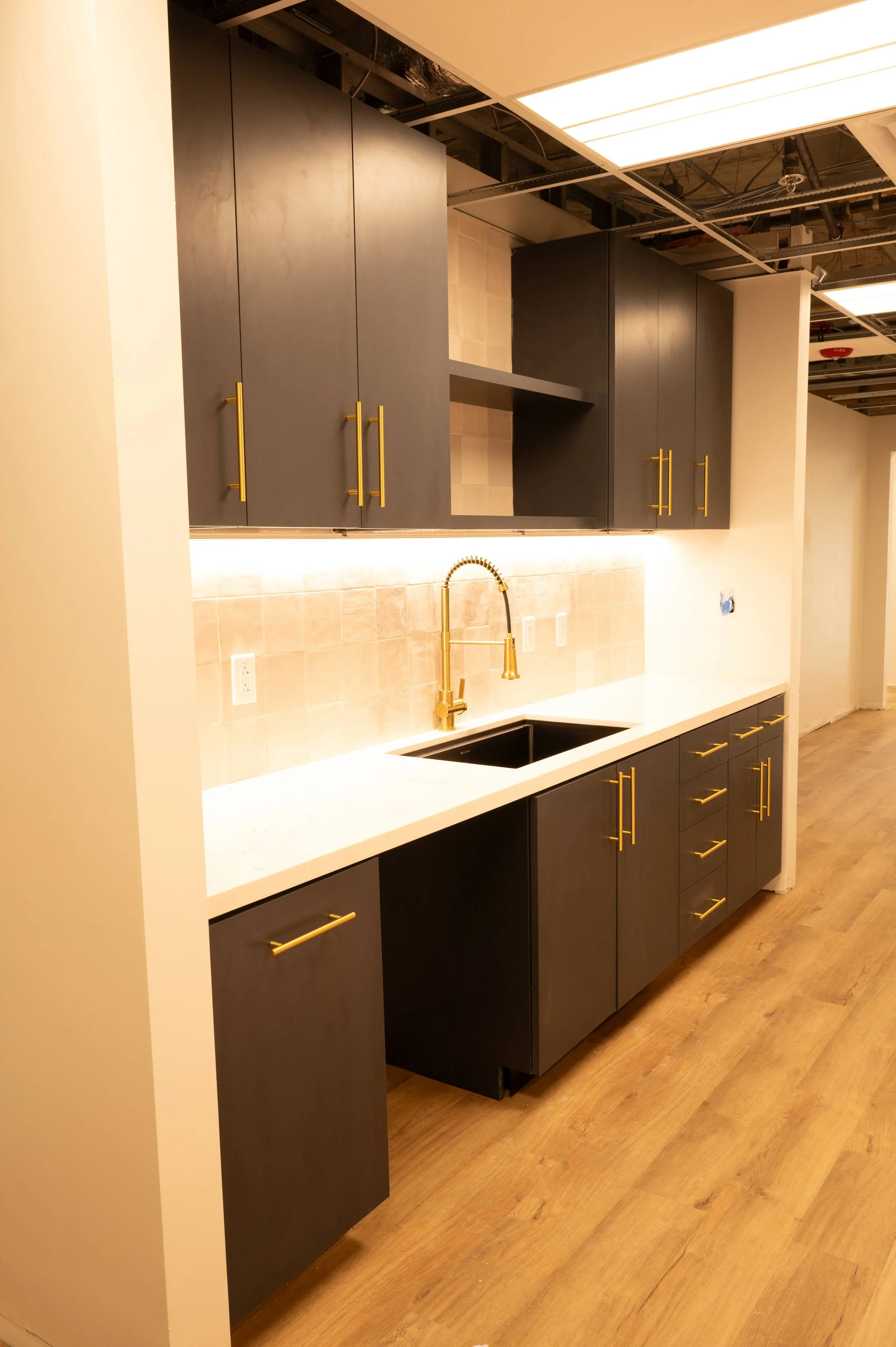 Modern kitchen with blue cabinets and gold handles, a white countertop, black sink, gold faucet, beige tile backsplash, and exposed ceiling with lighting.